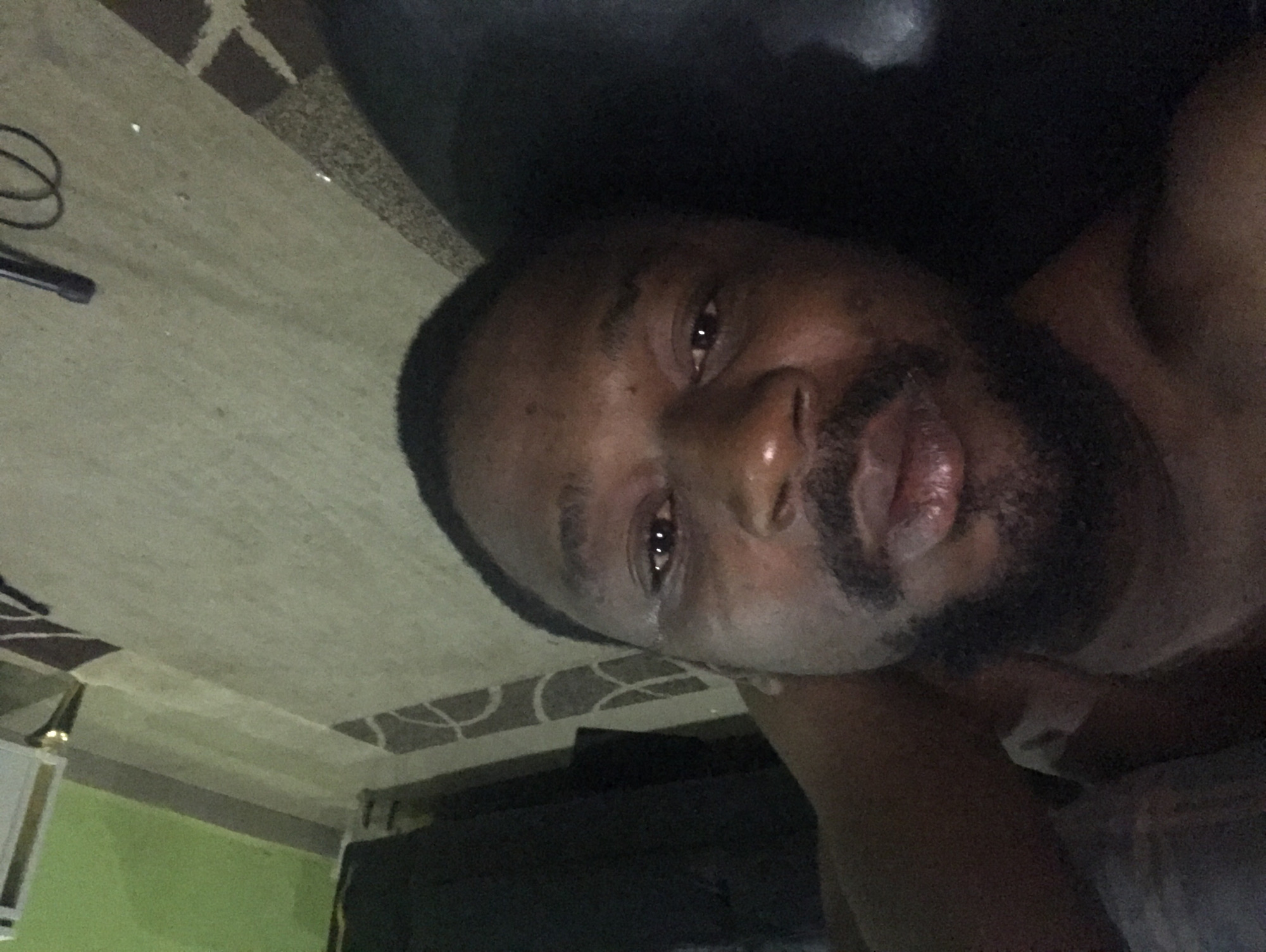 The image displays a close-up, slightly angled portrait of a man with dark skin, short dark hair, and a neatly trimmed beard. He is looking directly into the camera with a neutral to contemplative expression. His upper body is visible, suggesting he is either shirtless or wearing dark clothing that blends with the dimly lit background. Reflections are visible in his eyes, indicating a light source.
The setting appears to be indoors, likely a personal space such as a home in Abuja, Nigeria, as provided by the context. The background features a light-colored carpet or rug with a distinct brown geometric pattern, seen at an angle. To the lower left, a dark coiled cable or wire is visible on the carpet, possibly belonging to an electronic device or microphone. Portions of a green wall and a white object, possibly a piece of furniture or an appliance, are also discernible in the lower left quadrant. A dark, indistinct object, potentially a pillow or part of a couch, supports the man's head. The overall lighting is low, suggesting the photo was taken in the evening, at night, or in a room with subdued illumination. No visible text is present in the image.