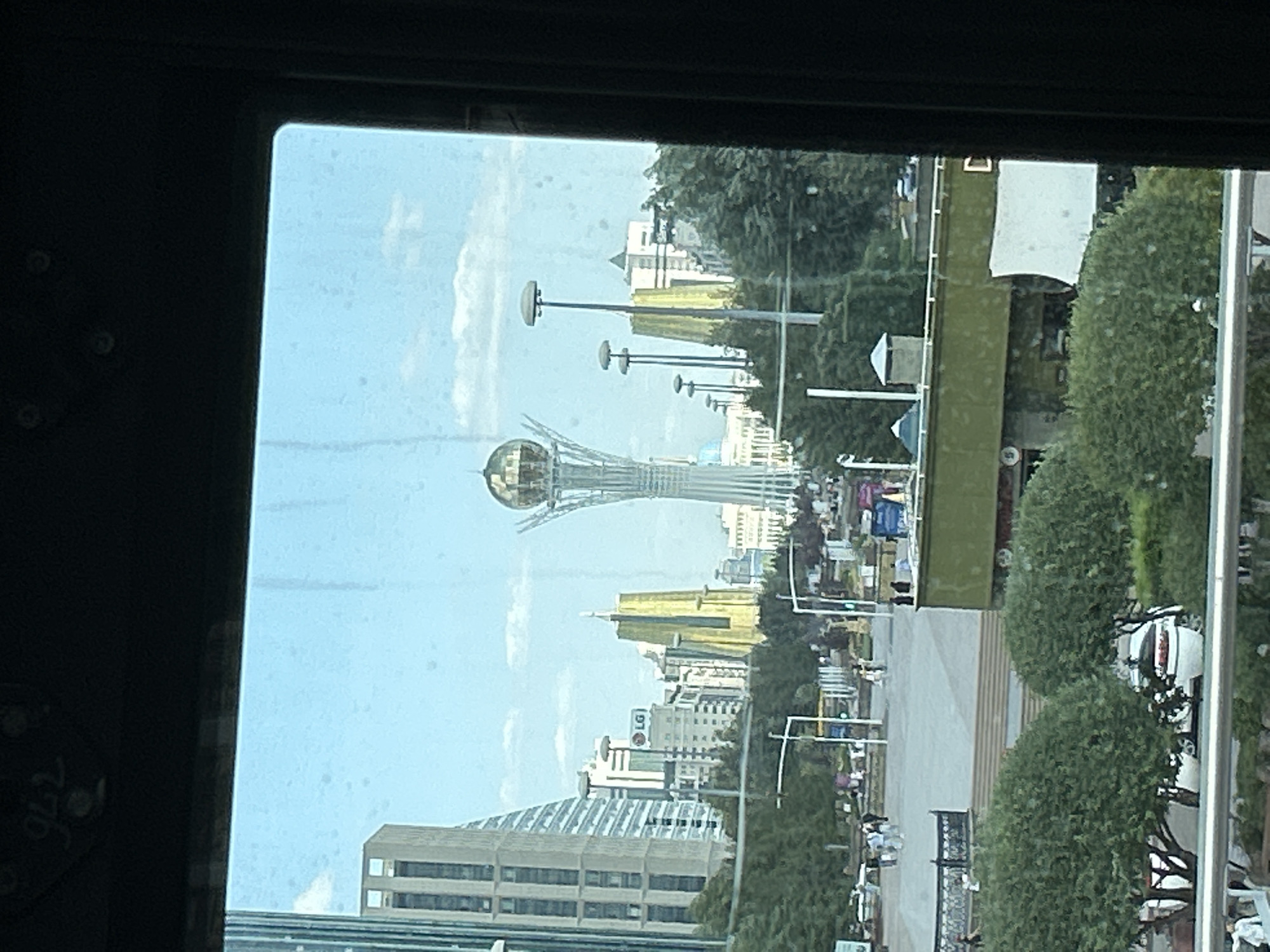 The image shows a view of the city of Astana, Kazakhstan, through a window, likely from a vehicle. In the foreground, a railing is visible, and beyond it, lush green trees and a white decorative planter with flowers. In the midground, there is a large open plaza with people walking and standing, lined with trees and streetlights. Buildings of various architectural styles are present, including modern skyscrapers and older structures, some with golden accents. A prominent landmark in the background is the Bayterek Tower, a distinctive observation tower topped with a golden sphere. The sky is blue with scattered clouds. The overall scene depicts an urban landscape on a clear day.