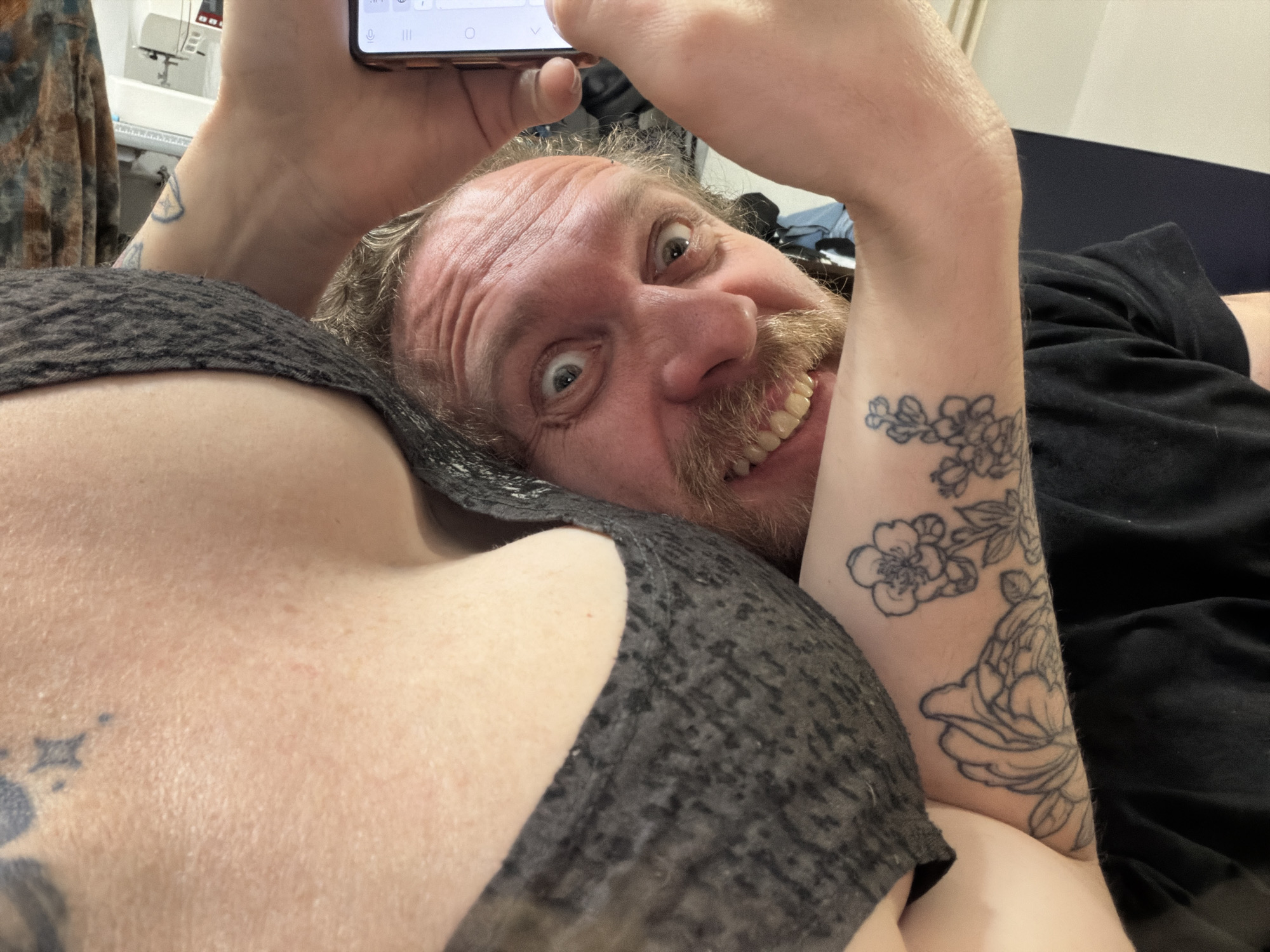 The image shows a selfie taken at a close angle. A person with fair skin, a reddish-brown beard, and short hair is looking directly at the camera with wide eyes and a smile, holding a smartphone above their head. The smartphone is black with a red trim. The person is lying on their back. In the foreground, another person's shoulder and chest are visible, covered by a gray and black patterned top. The arm of the person holding the phone has a floral tattoo on the inner forearm. A tattoo of a star-like shape is visible on the first person's shoulder. The background includes a glimpse of a dark blue or black bed or couch, a wall, and what appears to be a sewing machine on a table with various items around it. The location in the city of Saint Petersburg, Russia, is not discernible from the image.