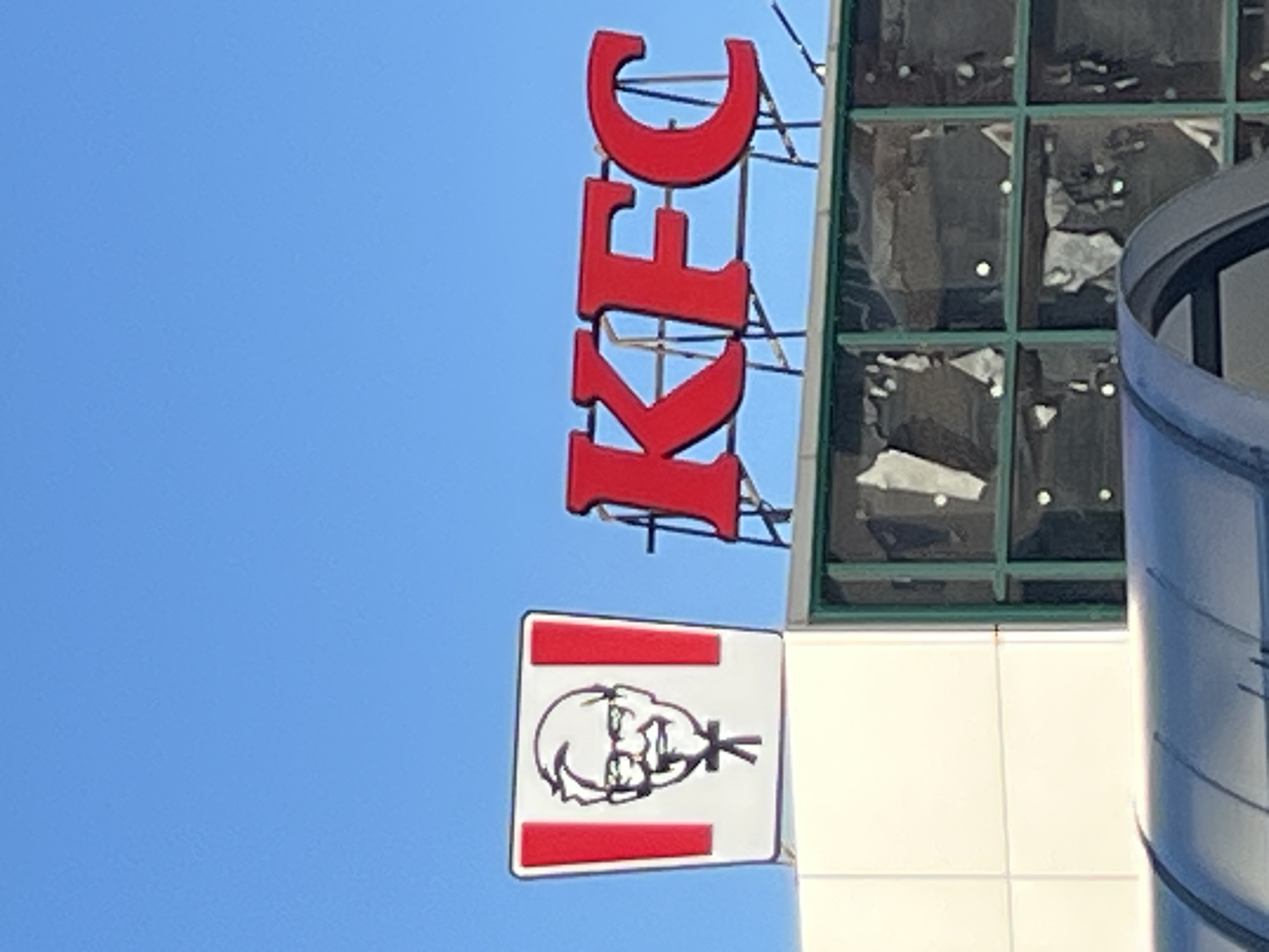 The image captures a sign for Kentucky Fried Chicken (KFC) located on a building in Minsk, Belarus. The sign features the iconic Colonel Sanders logo on the left, a square white panel with the Colonel's face, framed by red vertical stripes. To the right of this logo, the letters "KFC" are prominently displayed in large, red, three-dimensional lettering, mounted on a metal frame. The building itself has a white facade on the left, with a section featuring green-framed windows on the right. The windows appear to be broken or damaged, with large shards of glass missing. The sky above is a clear, bright blue. The overall scene is a commercial signage display against a backdrop of urban architecture under a clear sky.