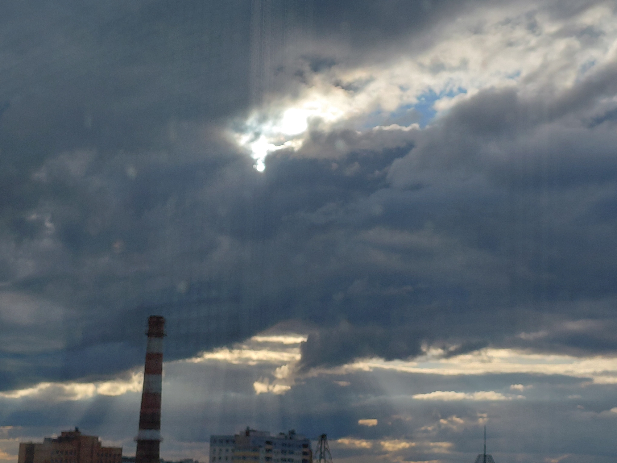 A wide-angle shot captures a cloudy sky with rays of sunlight breaking through. In the foreground, a tall, red and white striped smokestack stands prominently on the left side of the frame. To the left of the smokestack, a low-rise brick building is visible. On the right side of the frame, another building with multiple windows can be seen. The sky is dominated by dark, heavy clouds, with patches of bright white and blue visible where the sun is shining through. The image appears to be taken from a slightly elevated position, looking out over a cityscape. The overall mood is dramatic and somewhat ominous due to the dark clouds, but the sunlight offers a sense of hope. The location is the city of Verkhnyaya Podstepnovka, Russia.