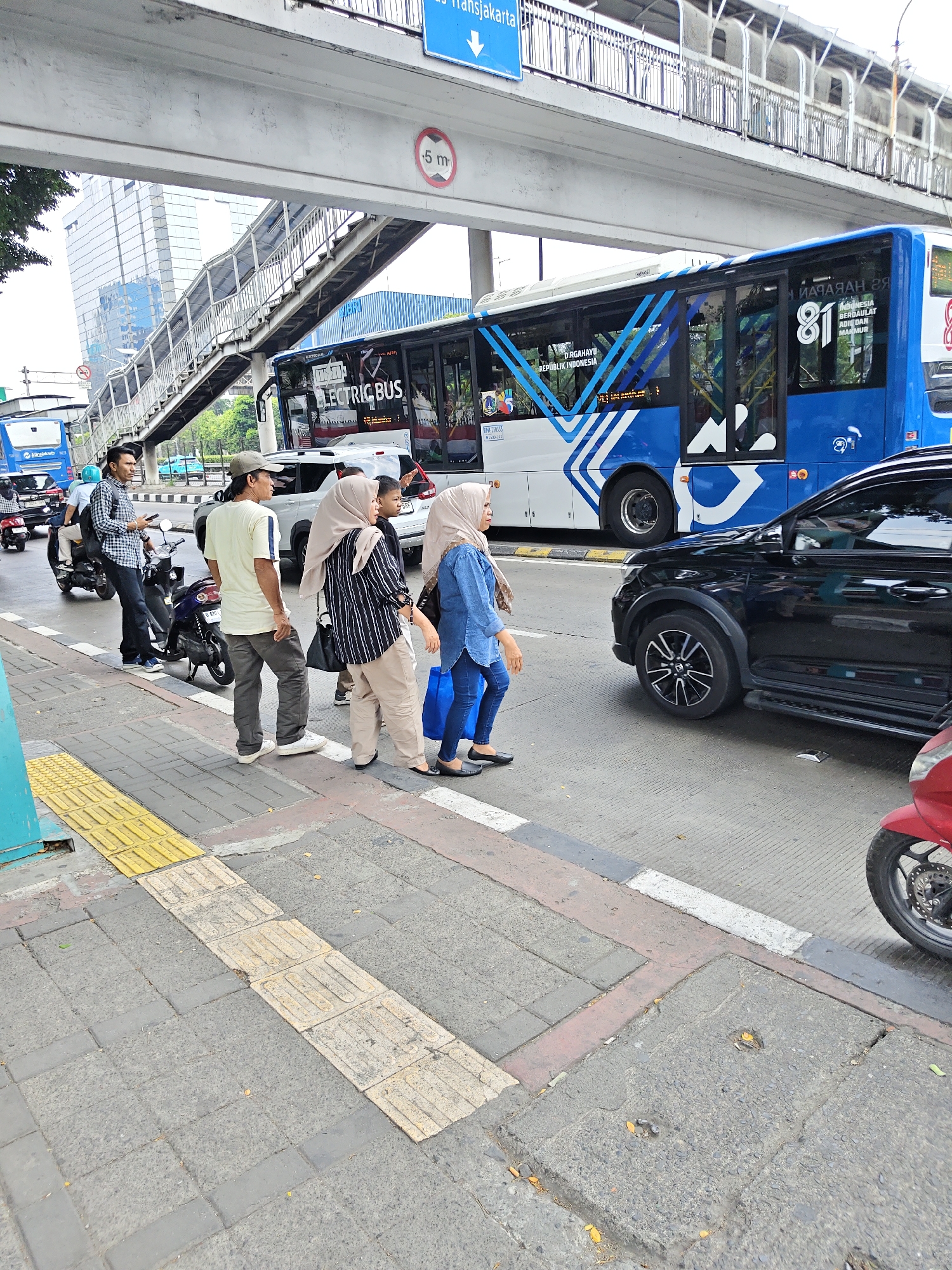 In West Jakarta, Indonesia, a group of people are gathered at a bus stop beneath an elevated pedestrian bridge. A blue and white electric bus, labeled "ELECTRIC BUS," is stopped, likely at the bus stop. Several individuals are waiting, some standing and others walking. One person is on a motorcycle, facing away from the camera. A black car is also present on the road. The scene takes place on a sidewalk with tactile paving, indicating a pedestrian-friendly area. The environment appears to be an urban setting with buildings and traffic.