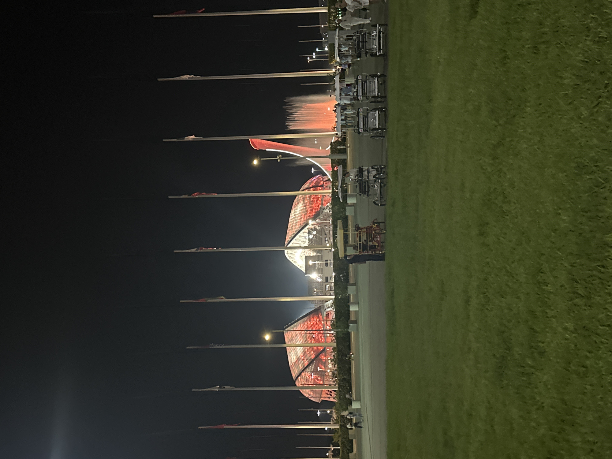 The image captures an exterior nighttime view of the Olympic Park in Sochi, Russia. The scene features illuminated architectural structures with distinctive geodesic dome-like roofs, glowing with red and white lights. Several tall flagpoles with flags stand prominently in the foreground. In the middle ground, a large fountain with red-lit water jets is visible. A row of golf carts or similar small electric vehicles is parked on a paved area. A few people are scattered throughout the scene, some walking and others near the vehicles. The foreground is dominated by a well-maintained grassy lawn.