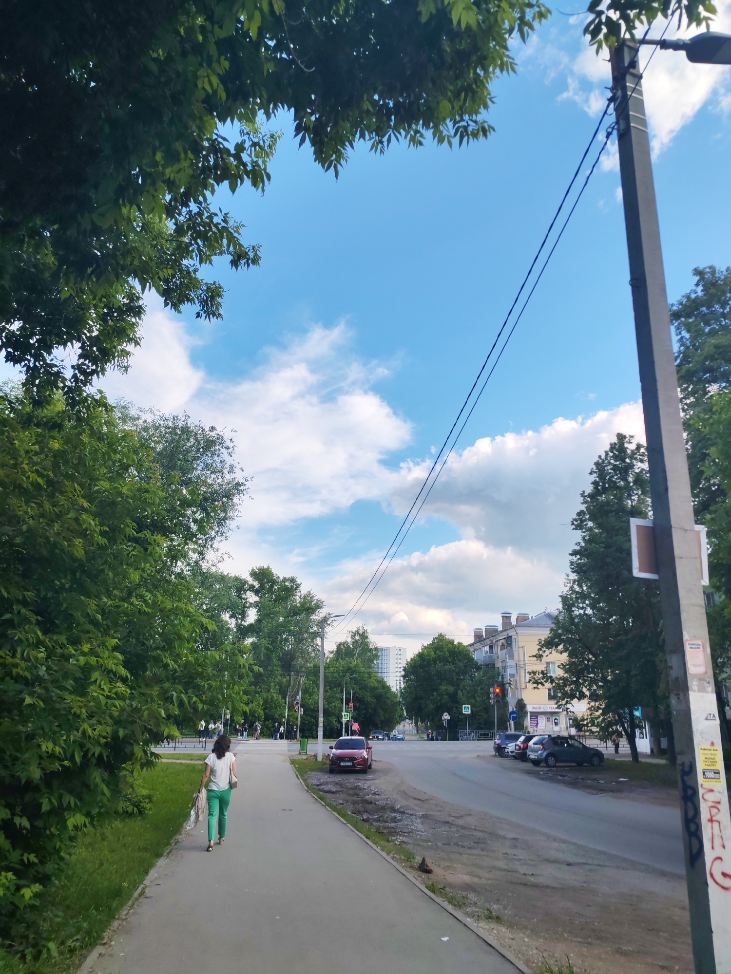 The image captures an urban street scene in Izhevsk, Russia, on a bright day with a clear blue sky and scattered white clouds. The perspective is from a paved sidewalk, looking down the street.
In the foreground, a woman with dark hair, wearing a white t-shirt and bright green pants, walks away from the viewer on the sidewalk. She is carrying a light-colored bag or item in her left hand. To her left, a dense line of lush green trees and bushes flanks the sidewalk, separating it from a grassy area.
To the right of the sidewalk, a road runs parallel, with several cars parked along its edge and some vehicles visible in motion further down. The road surface appears somewhat worn, with patches of dirt and gravel near the curb. A tall, gray utility pole stands prominently on the right side of the frame, with power lines extending across the sky. The pole has several small, affixed notices and some red graffiti, including what appears to be the letter "G" and numerical text that reads "1000 руб." (1000 rubles).
In the mid-ground and background, buildings of varying heights and styles are visible. One prominent building has a yellow facade and several red-capped chimneys. A sign on one of the buildings across the street partially displays the Cyrillic text "МОЙ ПАР", likely part of a business name. A red traffic light and a pedestrian crossing sign (a white triangle with a black walking figure) are visible at an intersection further down the road, where other pedestrians can be seen. The abundant green foliage suggests a summer or late spring season.
