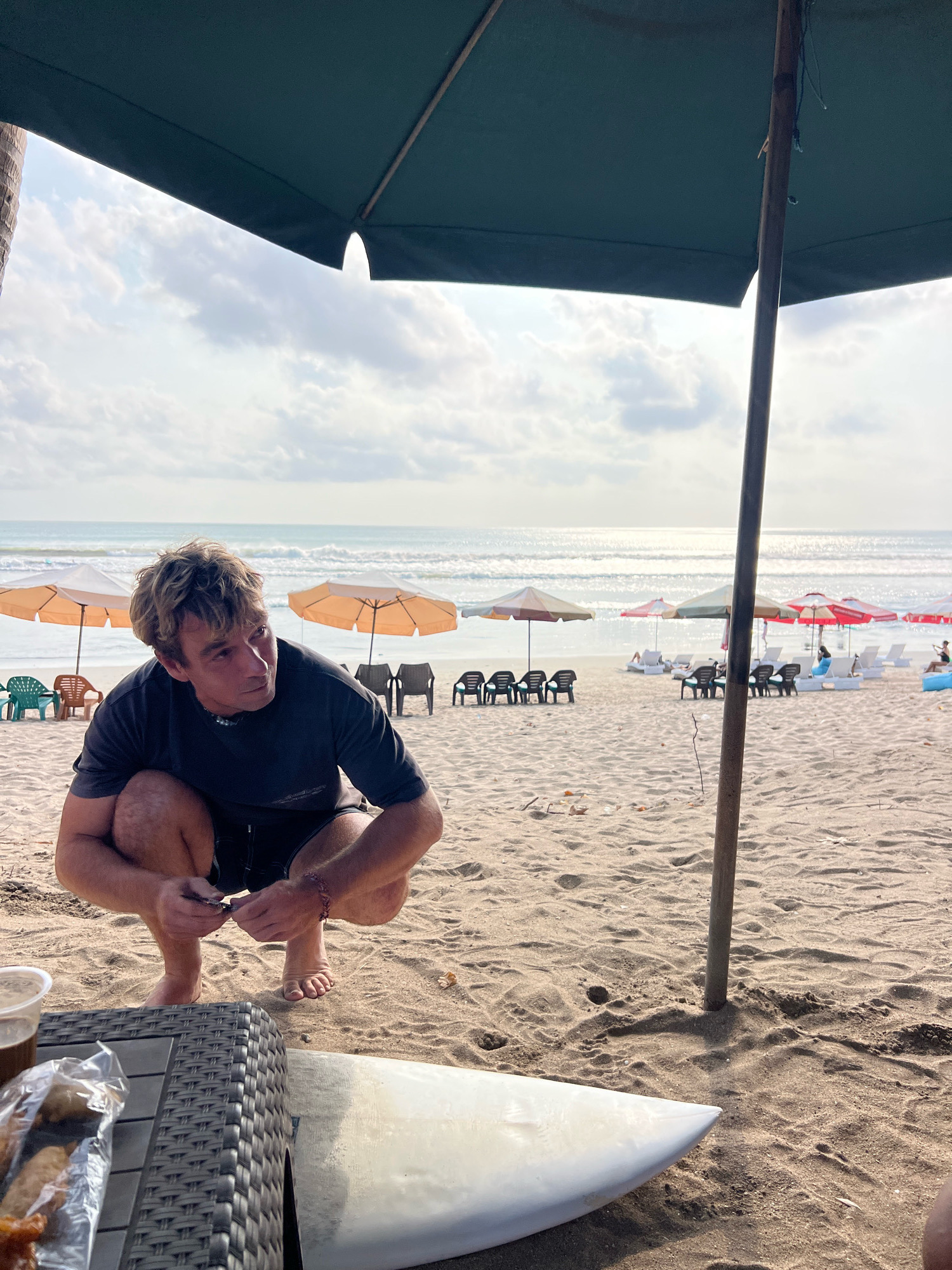 A man is crouching on a sandy beach in Kuta, Indonesia, looking to his left. He is wearing a dark t-shirt and shorts. In front of him, on a low-lying woven table, is a drink and a plastic bag of food. A white surfboard lies next to the table. The beach has several rows of lounge chairs and umbrellas, and the ocean is visible in the background with waves. The sky is cloudy with the sun shining through.