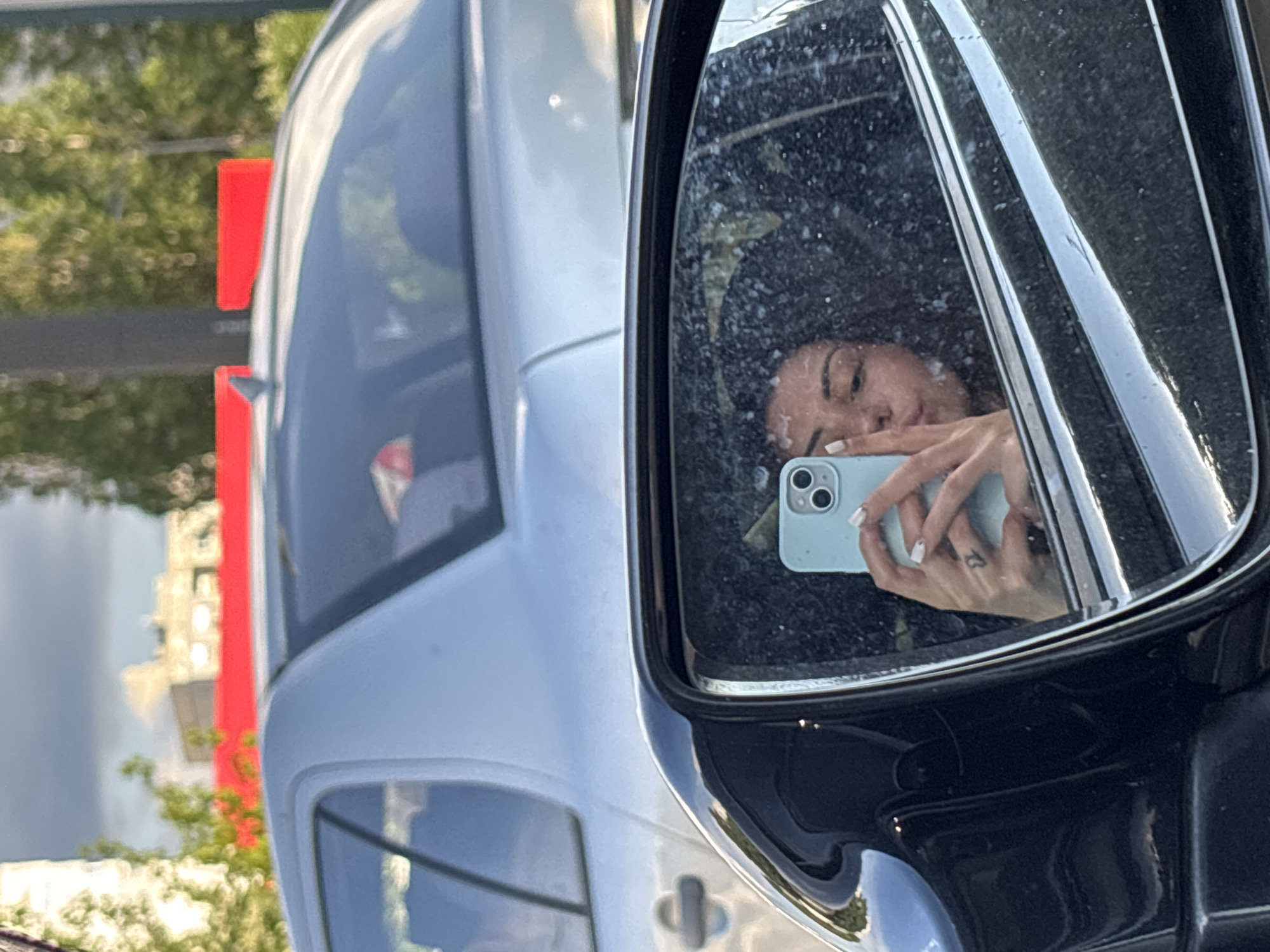The image is a selfie taken in a car's side-view mirror. The mirror is dirty, with many water spots or smudges. The subject is a young woman with dark, curly hair, looking at her phone. She is holding a light blue smartphone in her left hand, with her right hand partially visible. Her lips are pursed, as if she is blowing a kiss. There is a small tattoo on her left hand. In the reflection of the mirror, other cars are visible, including a silver car in front and what appears to be a blurred background of buildings and trees. The image appears to have been taken outdoors in a city, potentially during daylight hours. Without additional context or visible landmarks, pinpointing the exact location within Sofia, Bulgaria, is not possible.