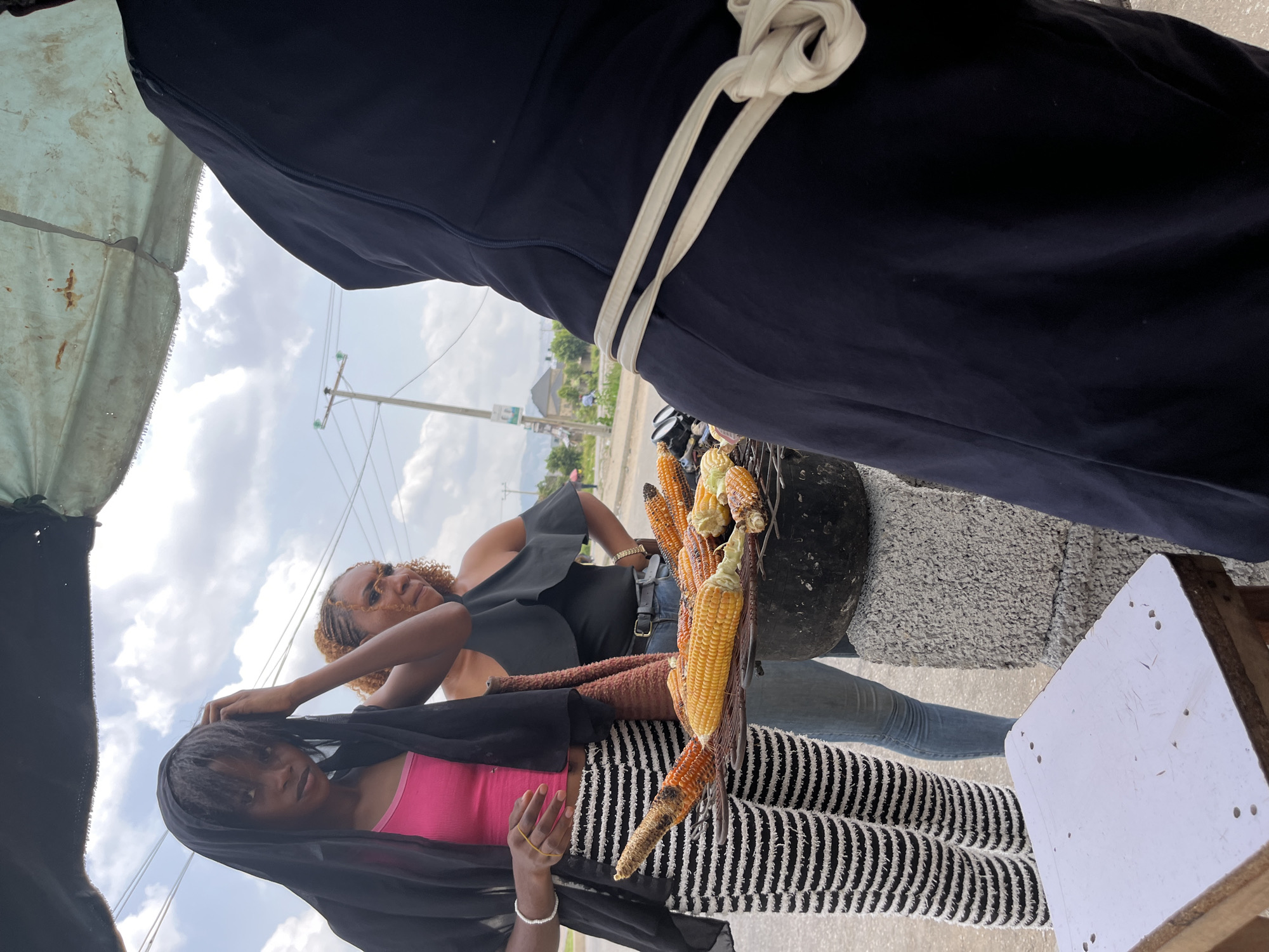 Two young women stand next to a vendor selling roasted corn on a street corner in Abuja, Nigeria. The vendor wears a navy blue garment secured by a white rope. A variety of roasted corn is displayed, some yellow and some darker. The woman closest to the camera is wearing a pink top, striped pants, and a black head covering. The second woman is wearing a black top and blue jeans. They appear to be interacting near the vendor's cart under an overcast sky. Power lines and a building are visible in the background.