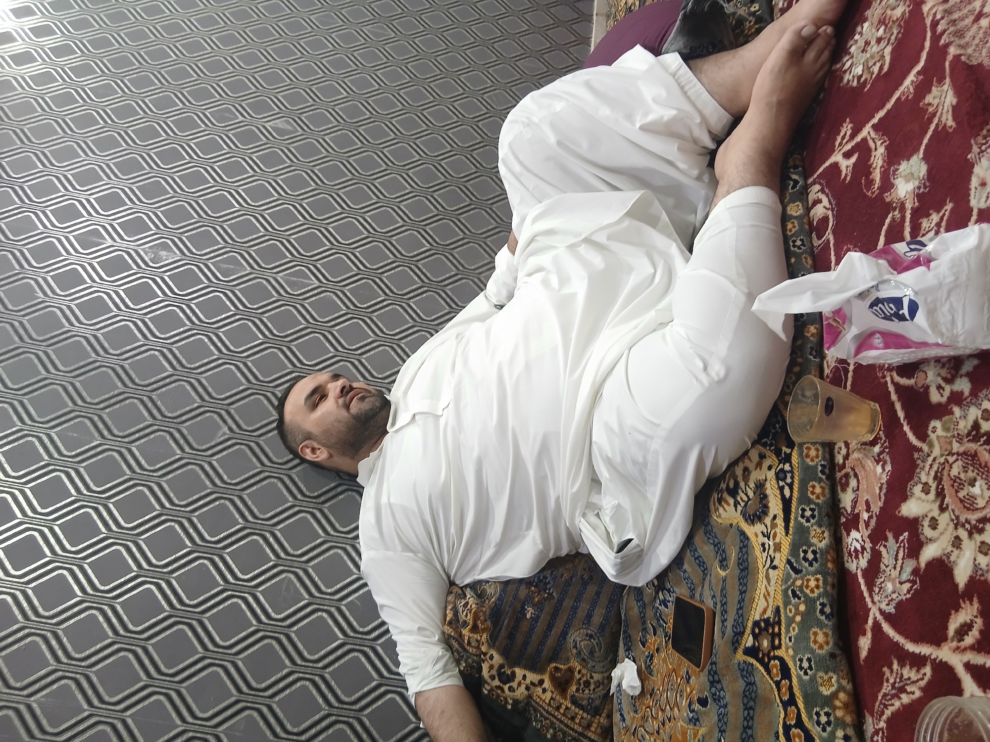 A man is lying on a carpet in Quetta City Tehsil, Pakistan. He is wearing a white thawb. His head is on a patterned gray surface. Next to him on the carpet is a glass, a tissue box, and a smartphone. The carpet is a mix of red, brown, and gold.