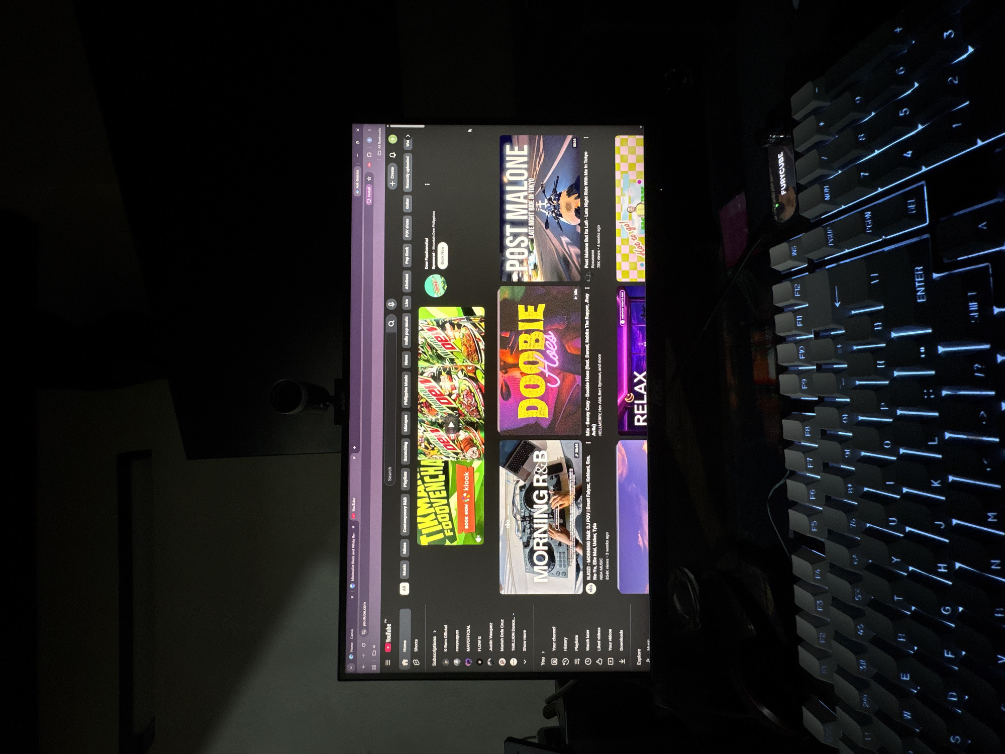 The image displays a computer monitor showing the YouTube homepage in a dimly lit room. The monitor is positioned in front of a keyboard with blue backlighting. On the screen, various video thumbnails are visible, including one sponsored by "Dew Foodvvencha!" and others featuring music artists and genres. A webcam is mounted on top of the monitor. The room appears to be a personal workspace or entertainment area. The presence of a keyboard with specific key legends like "F1" through "F12" and other standard keys indicates a functional computer setup. The overall lighting suggests it is nighttime or the room's ambient light is very low. The provided crops highlight specific areas of the screen and the keyboard. There is no indication or visual information present in the image that would suggest its location in Davao City, Philippines.