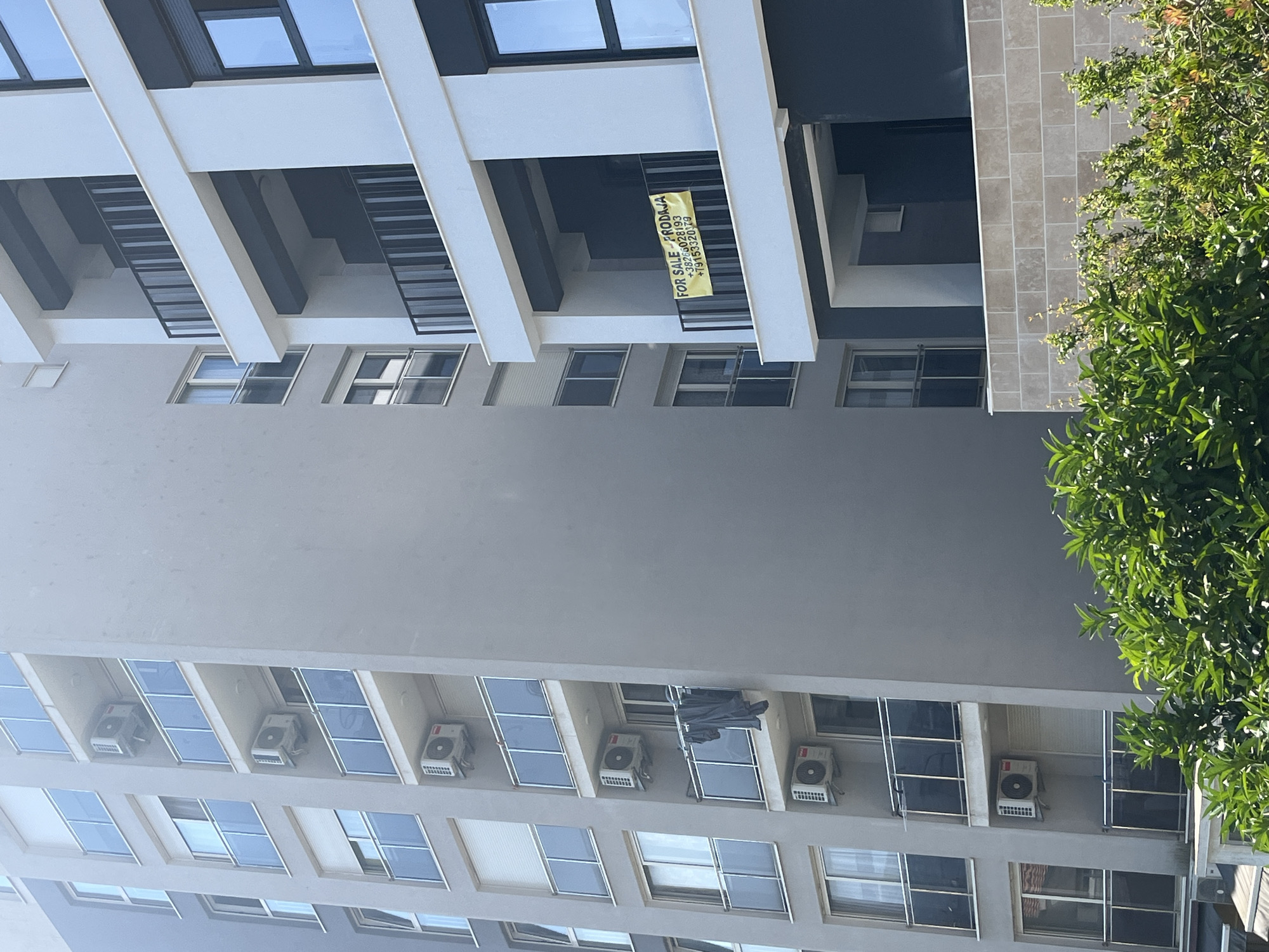 The image displays a modern apartment building in Budva, Montenegro, on a sunny day. The building features multiple balconies with glass railings and dark gray trim. On one balcony, a yellow "FOR SALE - PRODAJA" sign is visible, along with a phone number. Several air conditioning units are mounted on the exterior walls of the building. In the foreground, lush green foliage partially obscures the lower portion of the building. The sky is clear and bright blue.