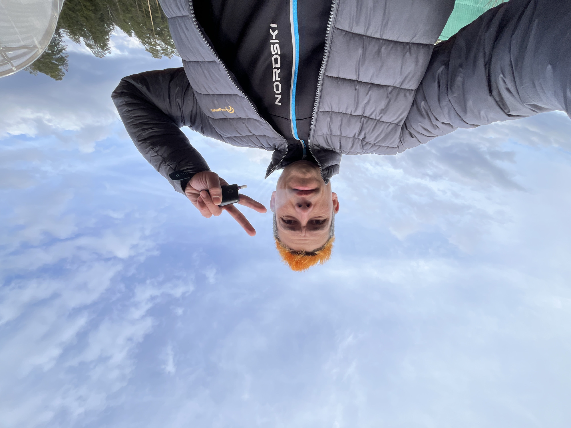 A selfie, taken from a low angle and then rotated, shows a man in front of a sky with clouds. He is wearing a black and gray puffer jacket over a black sweatshirt with the word "NORDSKI" in white and blue on the front. He has short orange hair. His left hand is raised, holding a small black object and making a peace sign. In the background, there is a blurred outline of trees and what appears to be a white structure. The location is in Chaikovsky, Russia.