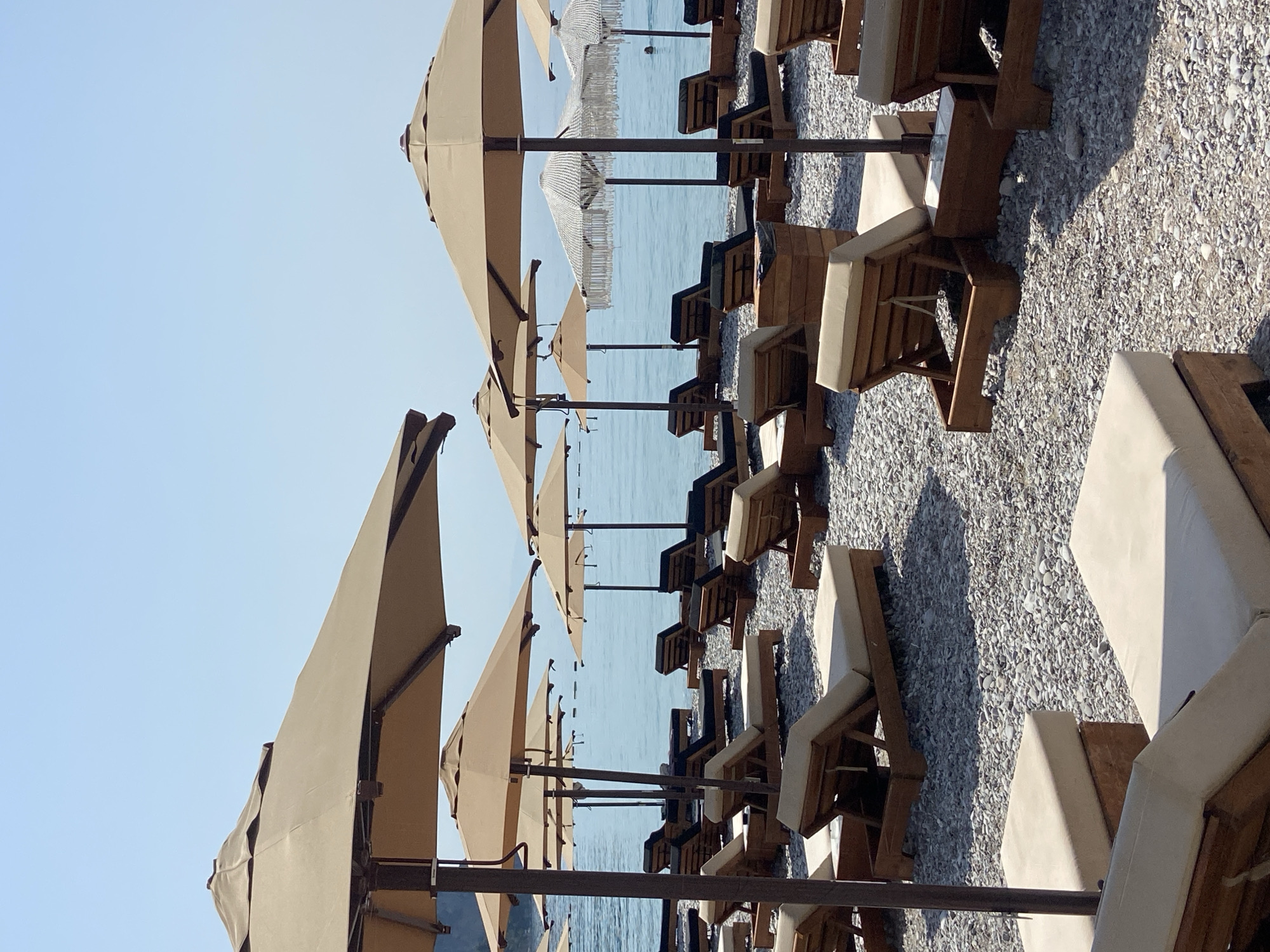 The image depicts a beach scene in Prijevor, Montenegro, characterized by a large expanse of pebbles. Numerous beach loungers, mostly made of wood with beige cushions, are arranged across the pebbled surface. Several large, tan-colored umbrellas provide shade for these loungers. The sea, a tranquil blue, is visible in the background, meeting a clear, bright sky. A few faint, horizontal lines are discernible on the water's surface, possibly indicating buoys or a distant coastline. The overall impression is one of a serene and prepared beach awaiting visitors.