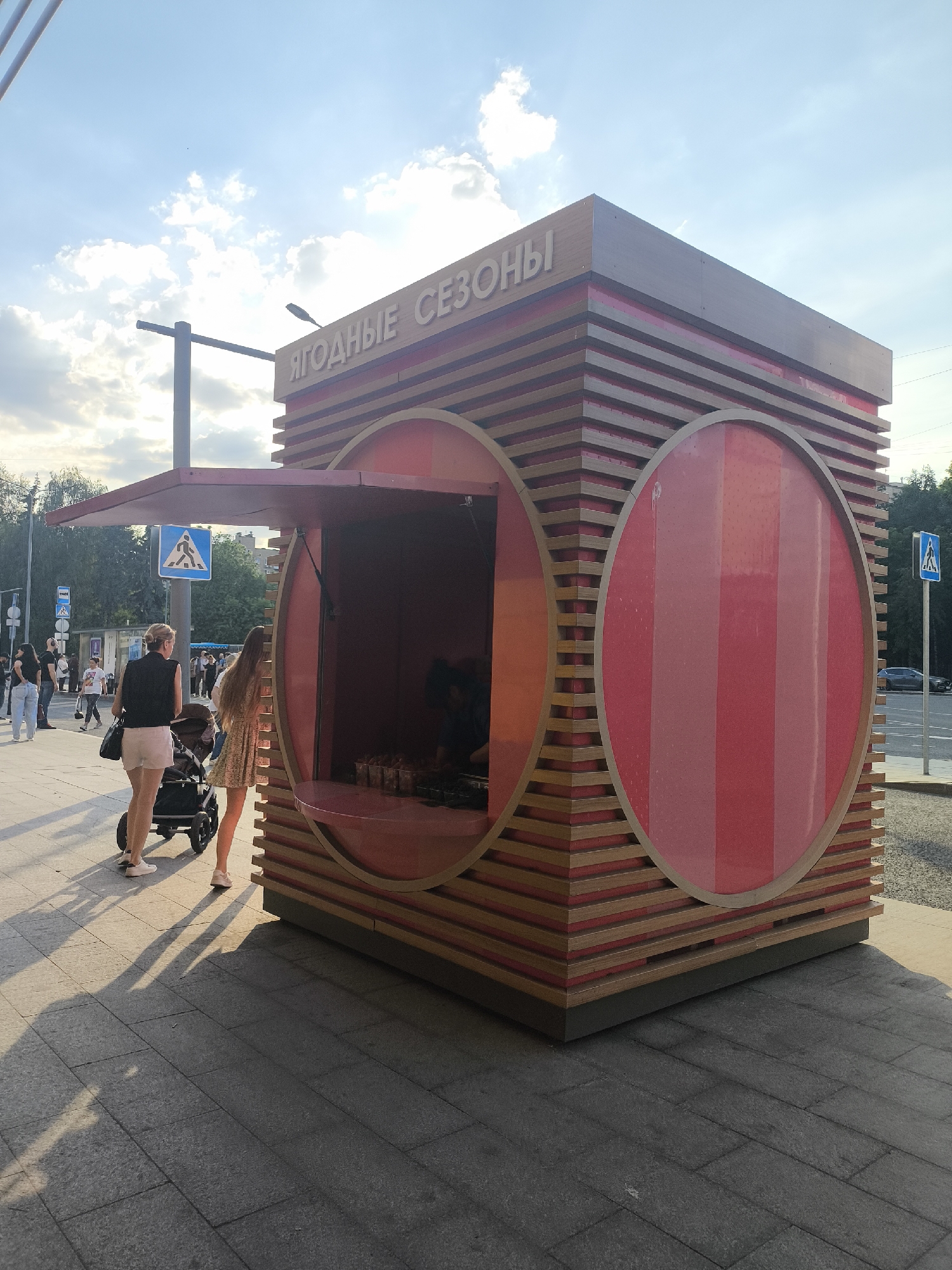 The image depicts an outdoor scene in an urban setting, likely in Unknown, Unknown. The focal point is a unique kiosk with a wooden structure and circular windows, adorned with the Russian text "ЯГОДНЫЕ СЕЗОНЫ" (YAGODNYE SEZONY), which translates to "Berry Seasons."
To the left, two women are visible, one pushing a stroller. People are walking in the background along the sidewalk. The environment includes sidewalks and trees. A pedestrian crossing sign is also visible.