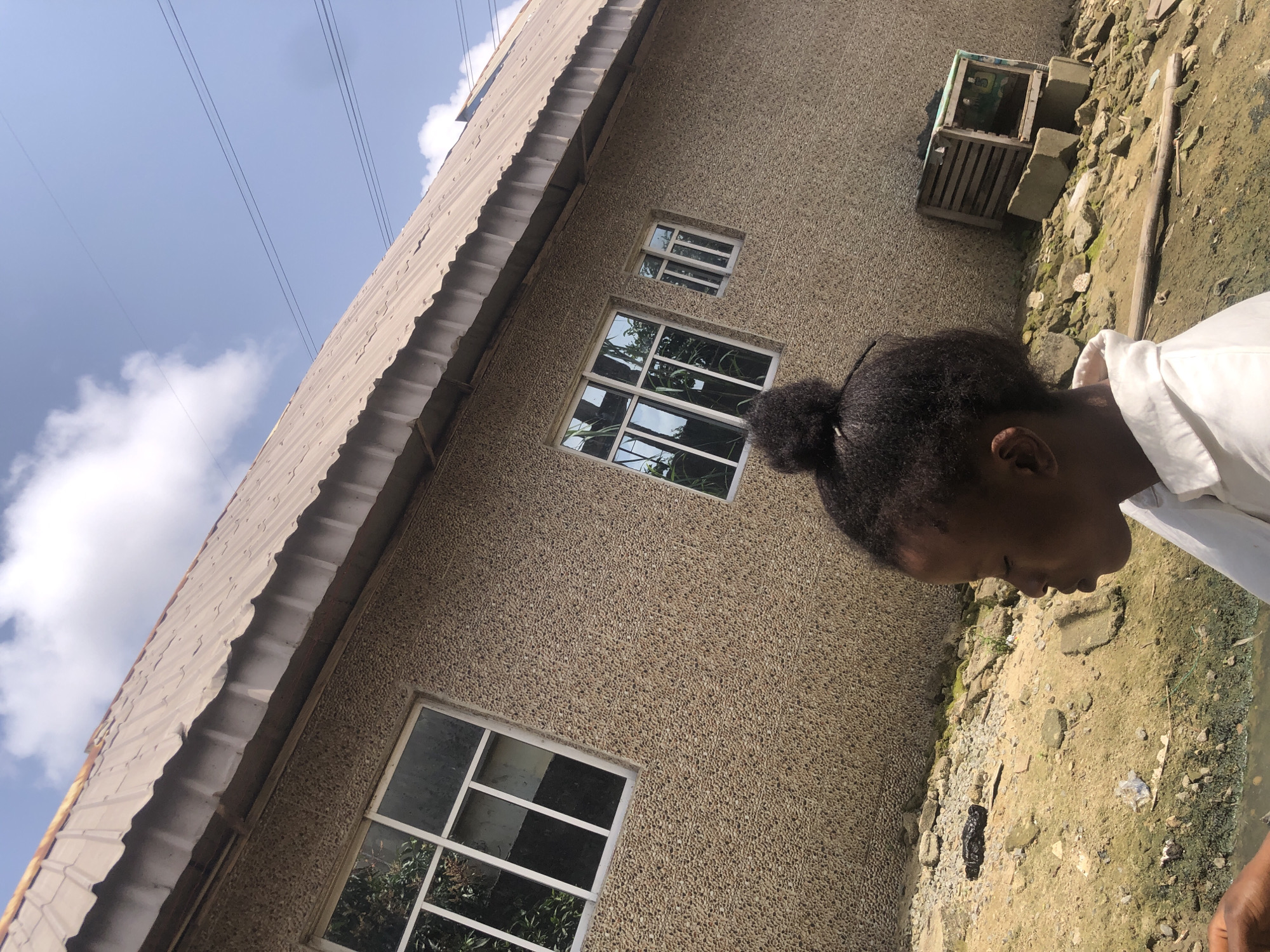 A young girl with her hair pulled into a bun is positioned in the foreground, facing to the left and looking down. She is wearing a white collared shirt. The background shows the side of a building with a textured, pebble-dash finish and a corrugated metal roof. There are three windows visible on the building's wall. To the right of the building, there is a wooden crate on top of concrete blocks. The ground in front of the building is uneven and appears to be a mixture of dirt, rocks, and some debris, with a hint of standing water or mud. Power lines are visible in the sky above the building. The overall scene suggests an outdoor environment in Ado, Nigeria.
