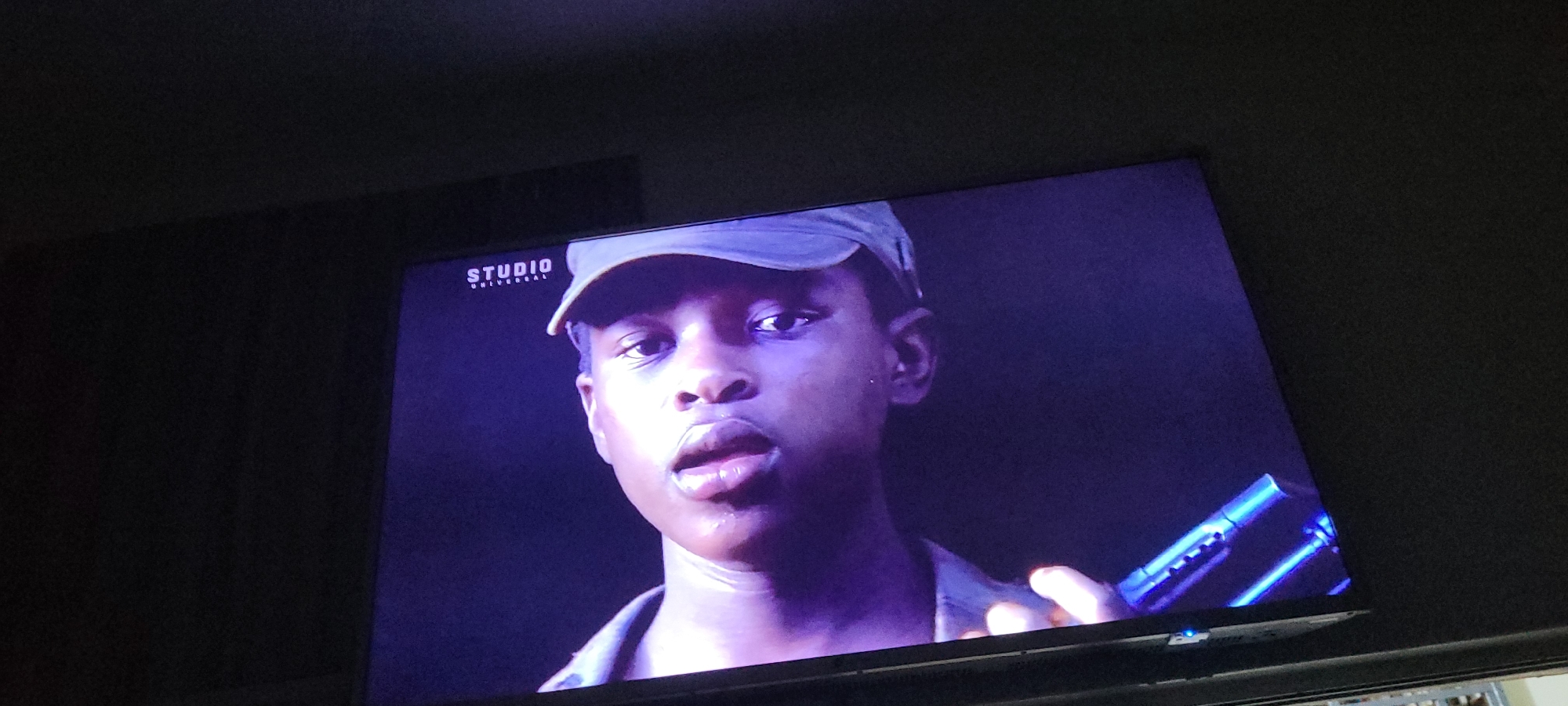 A close-up shot shows the face of a young person wearing a cap and a military-style uniform. The person's mouth is slightly open, and they appear to be speaking or reacting to something. In the lower right corner of the frame, a blue object, possibly a firearm or a piece of equipment, is visible. A "STUDIO UNIVERSAL" logo is present in the upper left corner of the screen. The background is dark and indistinct, suggesting an indoor setting or nighttime. The lighting is dramatic, casting shadows and highlighting the person's features. This image does not contain any information that would allow for its localization to Jos, Nigeria.