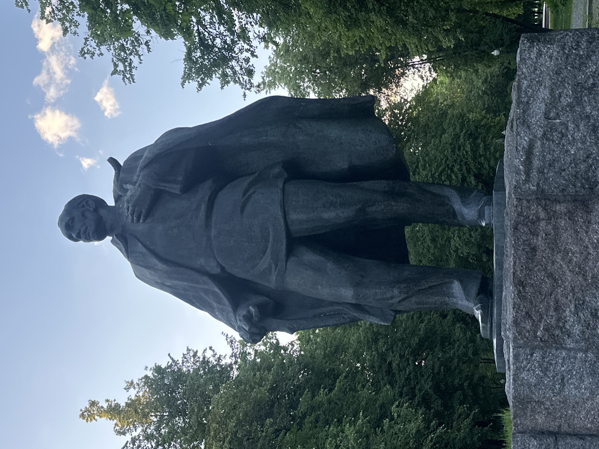 A large, dark statue of a man stands on a substantial, rough-hewn stone pedestal. The statue depicts a male figure wearing a long, flowing overcoat or cloak, an undershirt, and trousers. He appears to be looking straight ahead or slightly downwards, with a serious or contemplative expression. His left arm (viewer's right) is bent, holding what appears to be a cap or a rolled item against his chest. His right hand (viewer's left) is clenched into a fist, resting at his side or slightly forward.
The statue is situated outdoors, surrounded by dense, lush green foliage of trees, suggesting a park or a forested area. The background features a bright blue sky with scattered white, fluffy clouds. Sunlight is visible filtering through the tree canopy, particularly on the right side of the image, indicating either morning or late afternoon. The overall lighting suggests a clear day. No visible text is present on the statue or its pedestal. The scene is consistent with a public monument found in a city like Minsk, Belarus, where significant statues often commemorate historical figures.