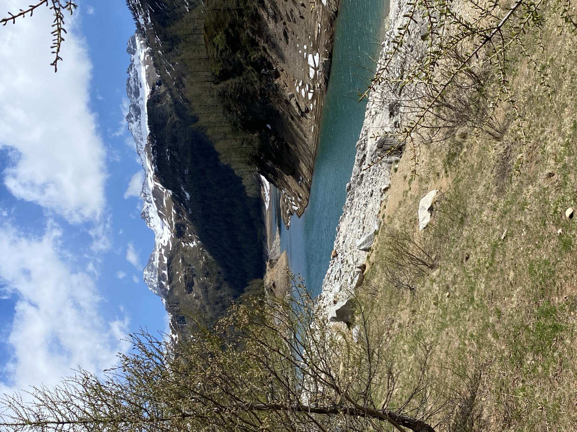 The image depicts a landscape in Baceno, Italy, featuring a lake or reservoir situated between steep mountains. The mountains are partially covered in snow, suggesting a high altitude or colder climate. A rock face or cliff runs along the side of the lake, transitioning into grassy terrain with sparse vegetation, including trees. The sky is blue with scattered clouds. The image is taken from a high vantage point.