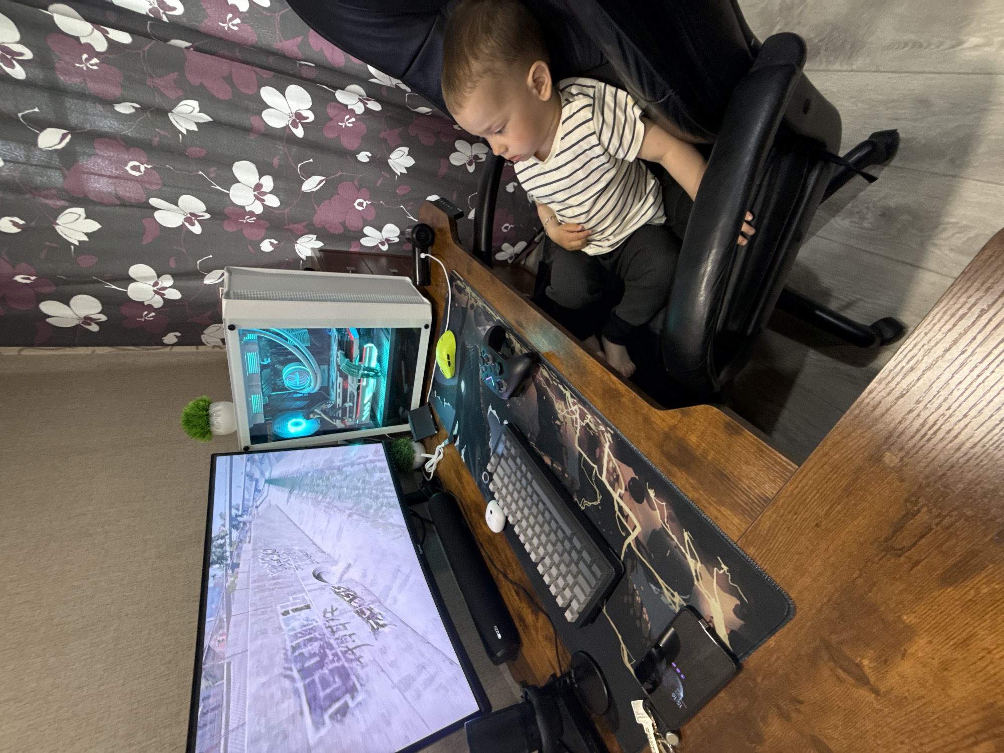 A young child sits in a black office chair at a wooden desk. The child is wearing a striped shirt and dark pants. The desk contains a computer monitor displaying a video game, a computer tower with illuminated internal components, a keyboard, a mouse, a microphone, and other accessories. A patterned curtain hangs in the background. The location is likely Rusanovo, Russia, based on the game displayed on the monitor, if accurately depicted.