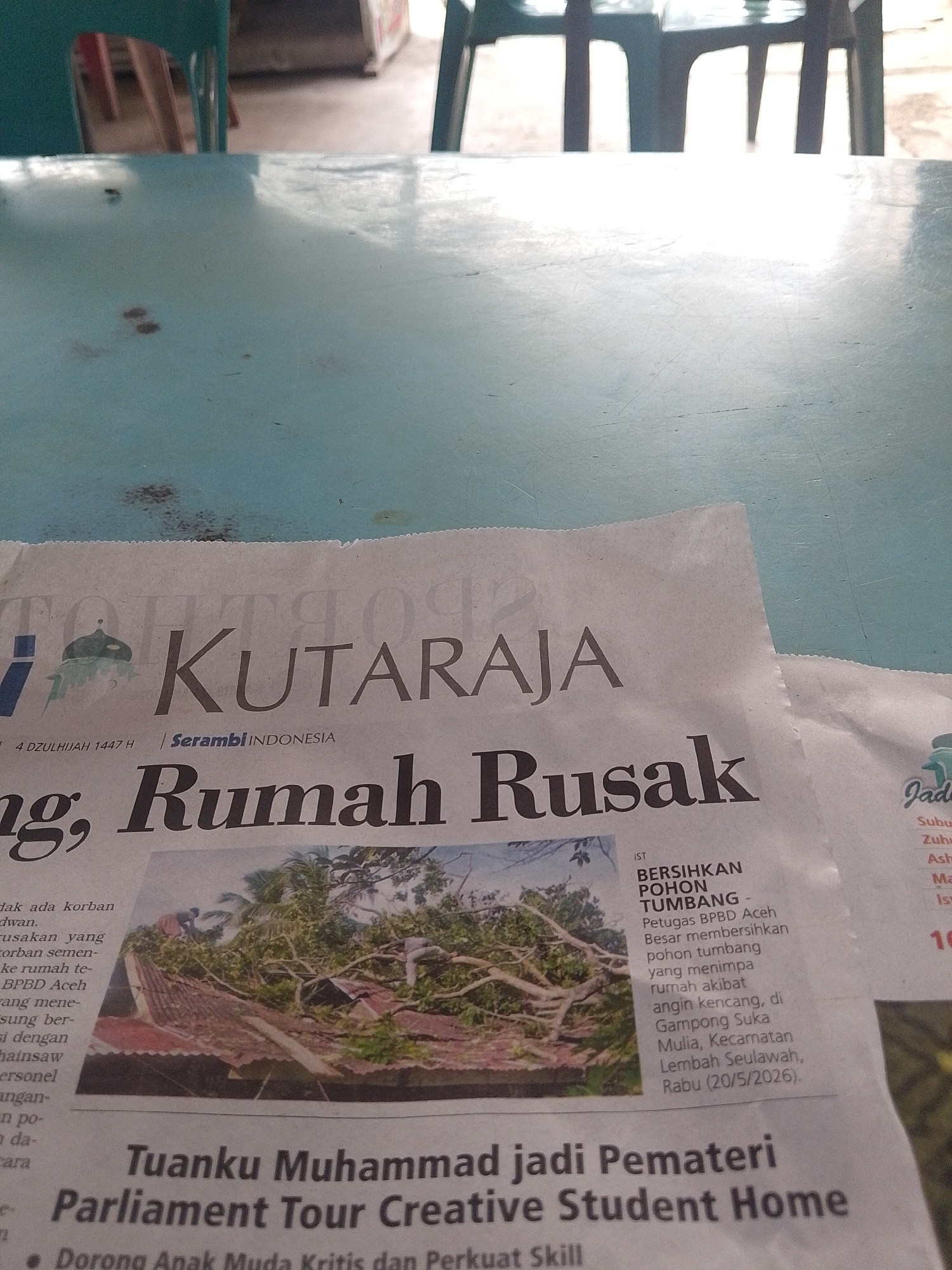 The image shows a portion of a newspaper on a table, headlined "KUTARAJA," with a subtitle "Serambi INDONESIA." The main headline below reads "Rumah Rusak," indicating damaged houses. An image shows people cleaning debris, likely from a damaged roof. Text adjacent to another picture reads "BERSIHKAN POHON TUMBANG - Petugas BPBD Aceh Besar membersihkan pohon tumbang yang menimpa rumah akibat angin kencang, di Gampong Suka Mulia, Kecamatan Lembah Seulawah, Rabu (20/5/2026)." Another headline states "Tuanku Muhammad jadi Pemateri Parliament Tour Creative Student Home." The newspaper snippet does not specifically mention the city of Lhokseumawe.