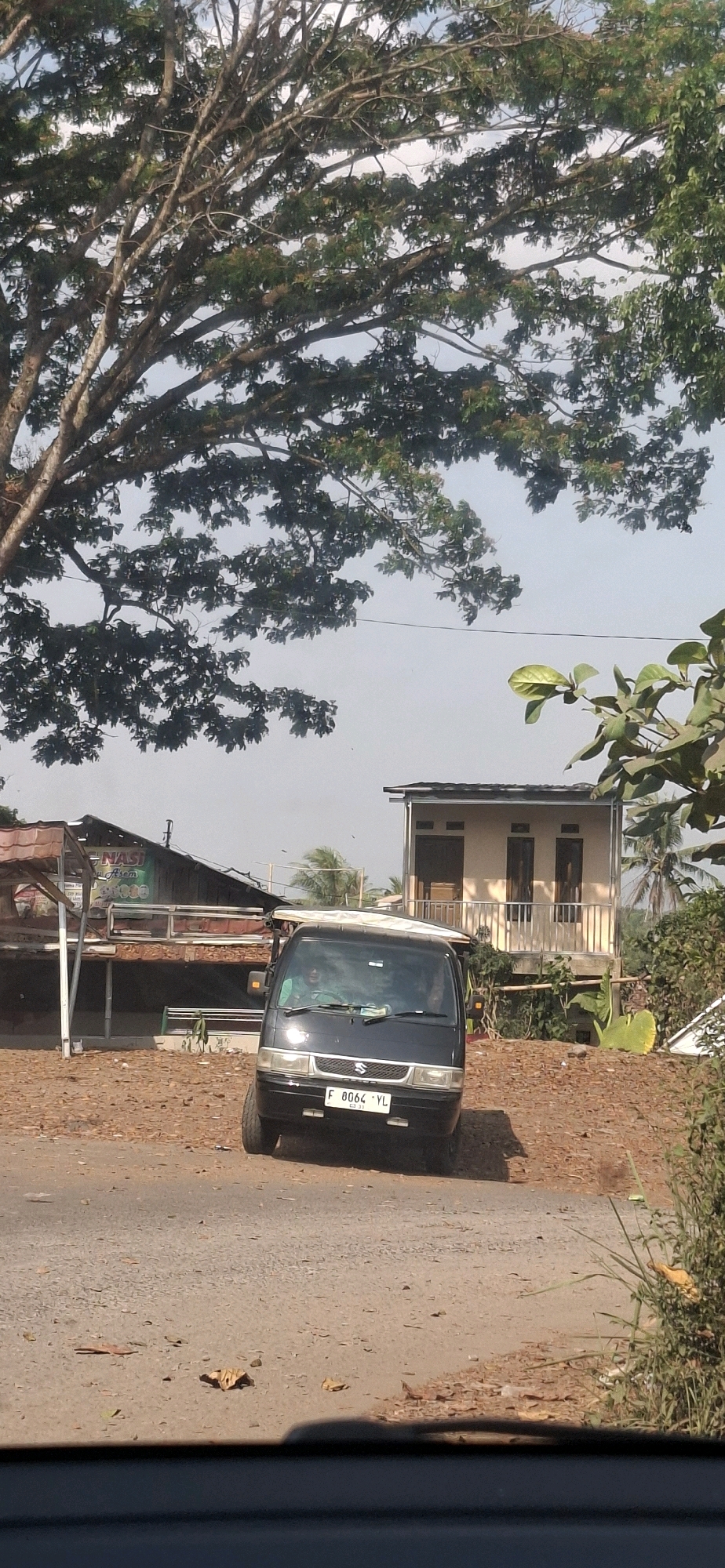 A black Suzuki Carry van is parked on a dirt road in front of a small, light-colored building with a balcony. A large tree with green leaves dominates the upper left portion of the image. To the left of the van, a covered food stall with a reddish-brown roof and brown wooden posts is visible. The ground is covered with dirt and fallen leaves. The sky is clear and light. The license plate of the van reads "F 8064 YL". Two people are visible inside the van, one in the driver's seat and another next to them. The scene appears to be in Cibinong Hilir, Indonesia.