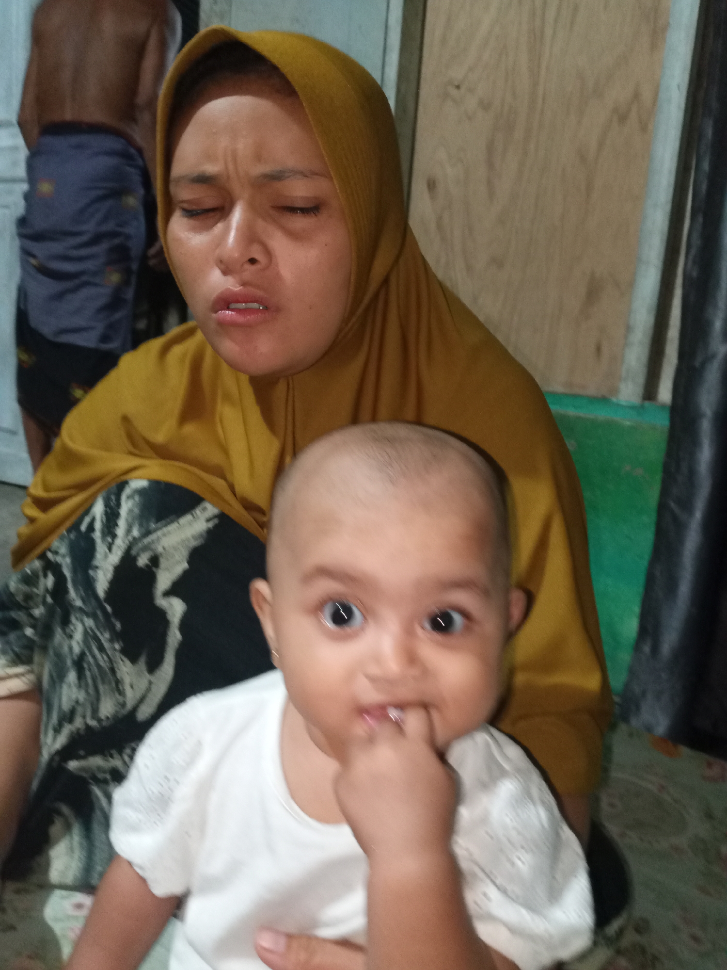 In a dimly lit, indoor environment, a woman wearing a mustard-colored hijab and a patterned skirt holds a baby. The woman's face, with eyes closed and mouth slightly open, conveys a strained expression. The baby, with wide-open eyes and a finger in its mouth, is dressed in a white top. A blurry figure of a person wearing a sarong is visible in the background. The room's features include a wooden wall and a dark curtain.