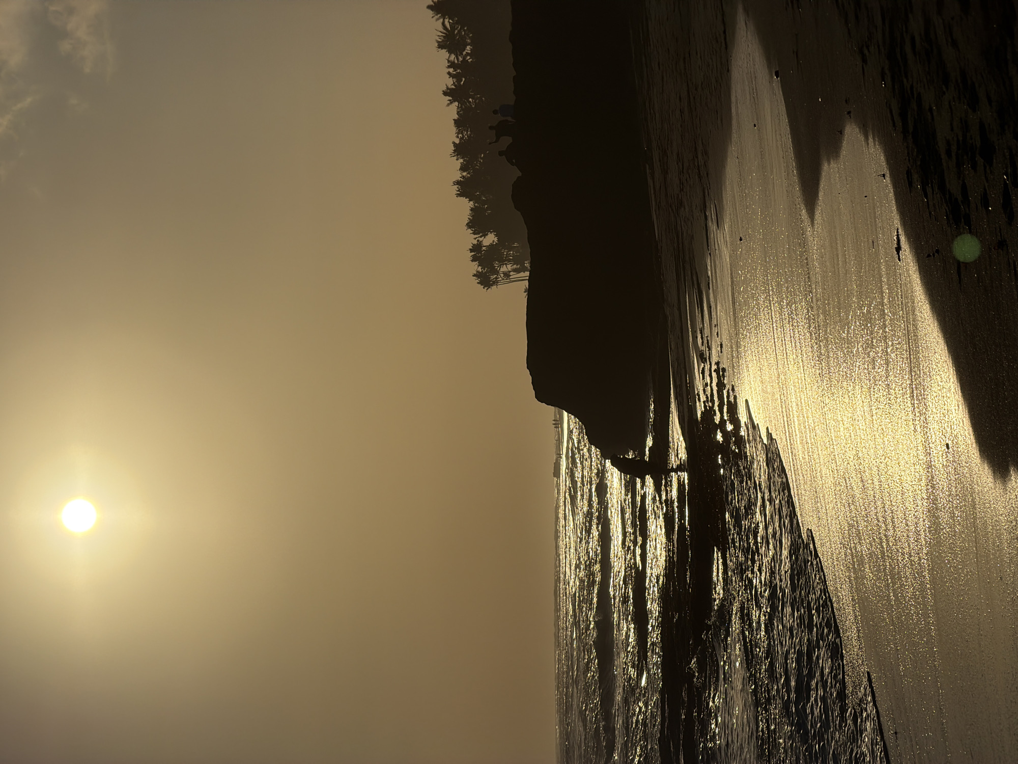 The image captures a sunset at Echo Beach in Canggu, Mengwi, Indonesia. The sun is low in the sky, casting a warm, golden light over the scene. The ocean is rough, with waves crashing against the shore and reflecting the sunlight. A person is silhouetted on the beach in the foreground, near the water's edge. Further down the beach, on a rocky outcrop, a group of people are gathered, also silhouetted against the sky. Palm trees line the top of the outcrop, forming a dark, dense silhouette against the hazy sky. The sand is wet and reflective near the water, with darker, dry sand further up the beach. The overall impression is one of a dramatic and serene sunset over a tropical coastline.