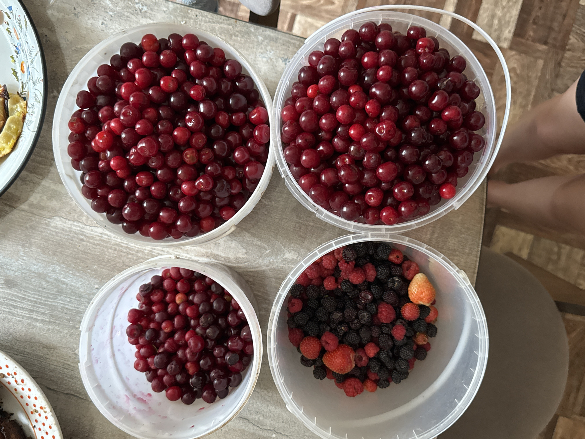 The image shows four clear plastic buckets filled with fruit. Two buckets are on the top row and two are on the bottom row. The top left bucket contains red cherries. The top right bucket is full of red cherries. The bottom left bucket contains a mix of blackberries, raspberries, and strawberries. The bottom right bucket is full of red cherries. The buckets are placed on a light-colored, textured surface. In the background, a portion of a plate with some food is visible. The image is taken from a high angle, looking down at the buckets. A person's legs and feet are visible at the bottom of the frame. The scene appears to be indoors. There is no information in the image to determine its location in Sharkan, Russia.