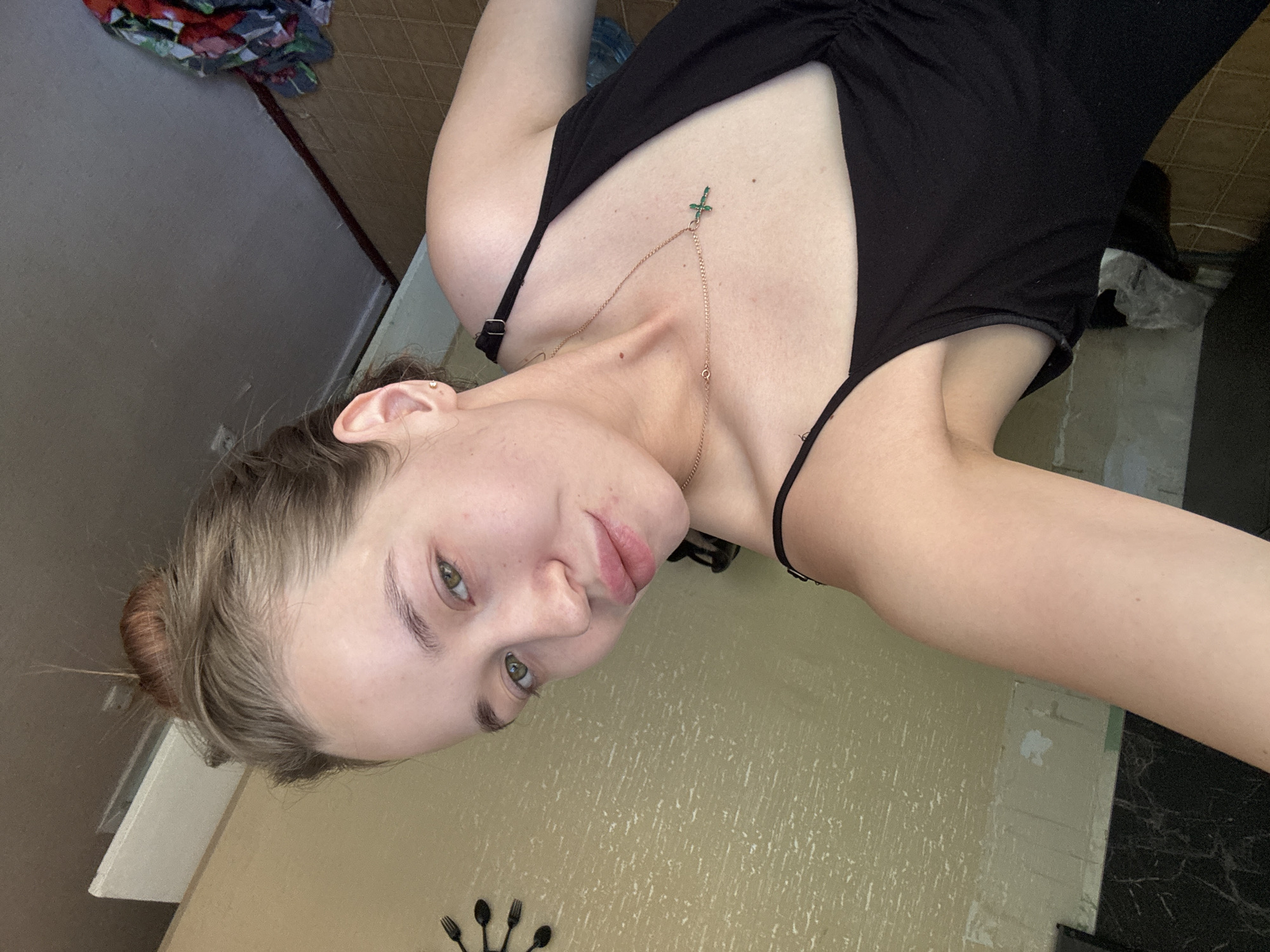 A selfie shot of a fair-skinned young woman, angled slightly towards the left. She has her hair in a bun. She is wearing a black, low-cut top with spaghetti straps and a gold chain necklace with a green cross-shaped pendant. The background consists of a beige-yellow textured wall. A set of utensils are visible on the wall in the corner. The location is unknown, including whether it is in the city of Chaikovsky, Russia.