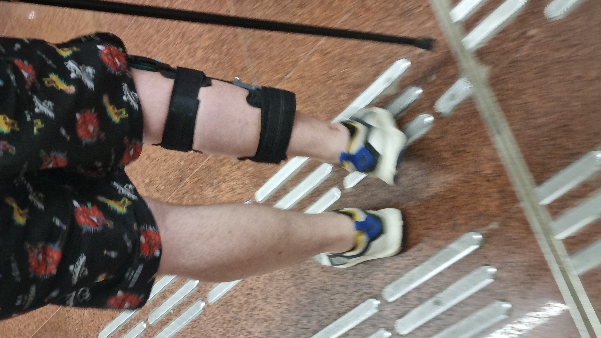A person is standing on a textured, reddish-brown floor with metallic strips embedded in it, possibly a tactile paving surface in a public indoor space. The person is wearing black shorts with a colorful pattern, a black knee brace, and white and blue athletic shoes. The person is holding a black cane. The image is a low-angle, full-body shot, focusing on the lower half of the person's body and the ground. There is no indication of the location being Moscow, Russia.