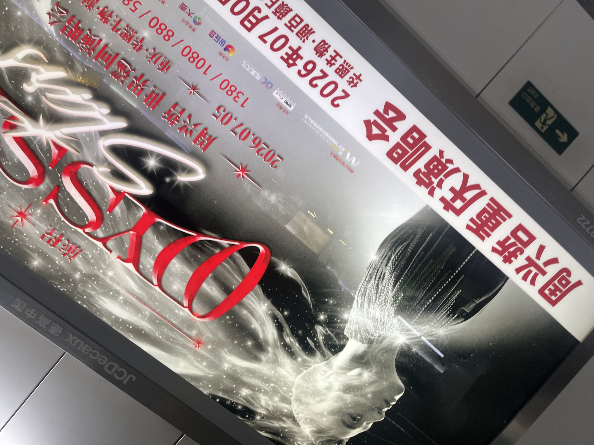 The image shows an advertisement billboard for a concert by "周兴哲" (Zhou Xingzhe) titled "Stars". The billboard is vertically oriented and angled upwards. The top left corner of the billboard has the "JCDecaux" logo along with Chinese text. The central focus of the billboard is the artist's name in large, stylized, glossy red and white lettering, reading "Stars". Below this, there is information about the concert, including the date "2026.07.05" and ticket prices "1380 / 1080 / 880 / 580". Further down, there is a large red Chinese text that appears to be part of the concert title. On the left side of the billboard, there is a black and white image of a person's profile with stylized white light emanating from their head and flowing into the background, resembling stardust. The bottom of the billboard contains more red Chinese text, which appears to be the event name "周兴哲重庆演唱会" (Zhou Xingzhe Chongqing Concert). A small green exit sign with an arrow pointing left and the word "EXIT" is visible in the bottom right corner of the scene, suggesting this advertisement is likely located in a public space like a subway station or an indoor venue in Chongqing, China. The city of Shapingba District, China is not explicitly mentioned on the billboard, but the presence of "Chongqing" in the concert title suggests a location within that municipality.