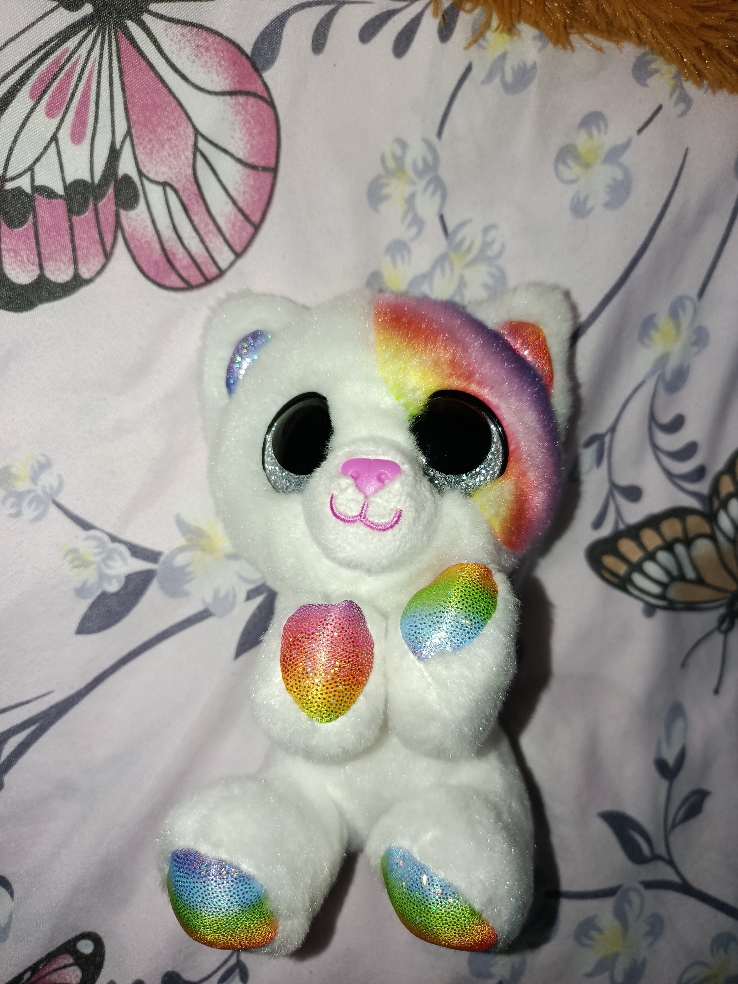 A white, fluffy stuffed cat toy with large, black, sparkly eyes and a pink nose is positioned centrally on a pink and white patterned fabric. The fabric features designs of butterflies and floral elements. The cat toy has a rainbow-colored patch over its left eye, extending to its ear, and rainbow-colored accents on its paws and ears. Its front paws are clasped together. The toy is illuminated by overhead lighting, casting a soft shadow to its right. The background is a continuation of the patterned fabric. The scene is an indoor setting, likely a domestic environment.