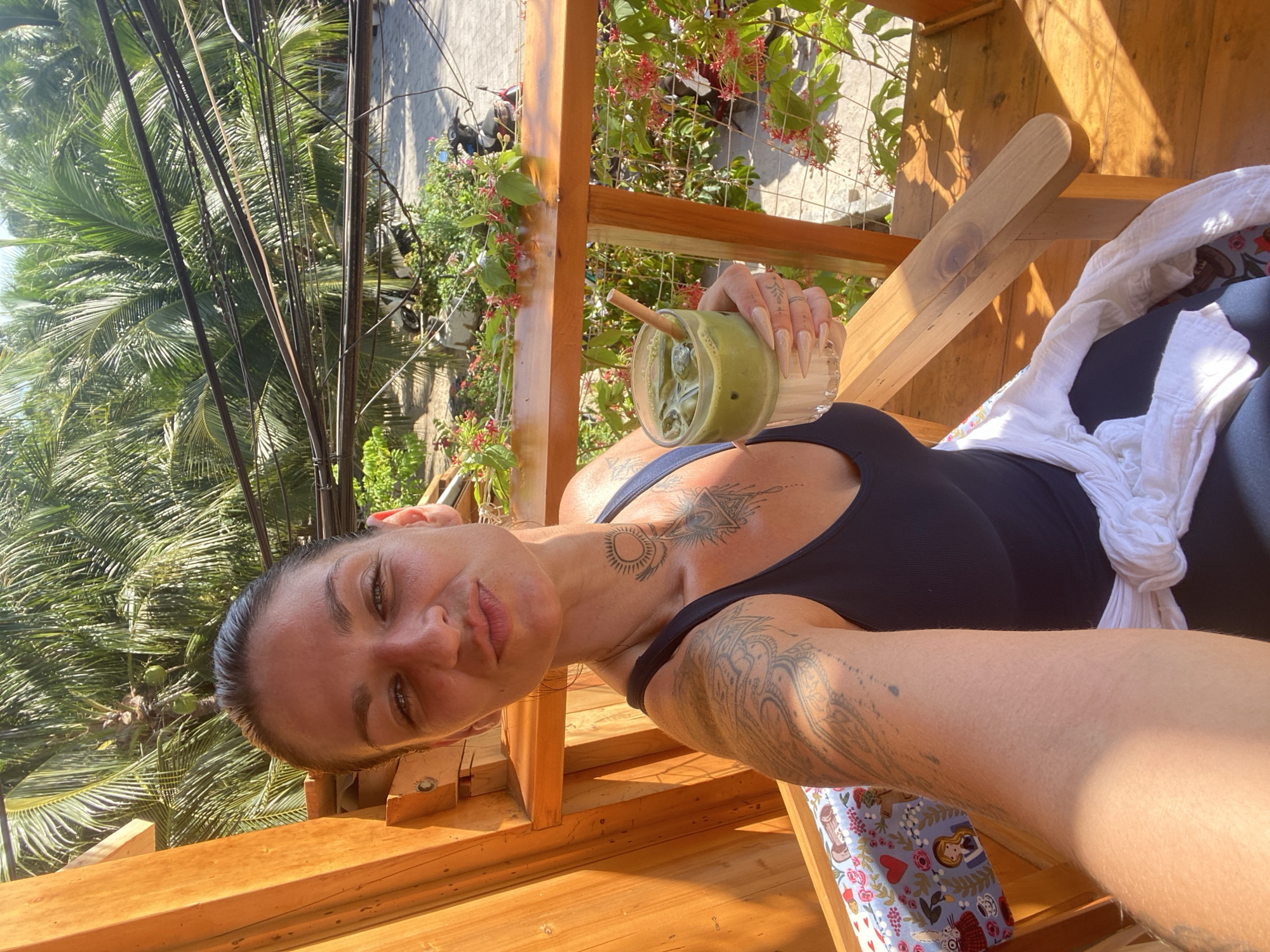 A woman with dark hair and tattoos on her neck and arms takes a selfie while sitting on a wooden chair on a balcony. She is holding a glass of a green beverage with ice and a straw. The background shows palm trees and electrical wires, suggesting an outdoor tropical setting. The image appears to be taken during the day. There are no visible signs indicating the specific location within Phu Thuy Ward, Vietnam.