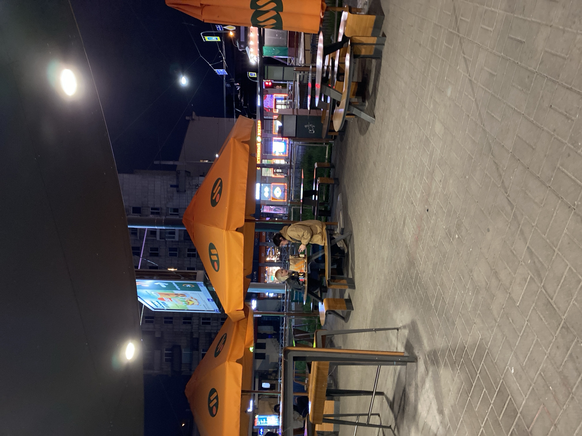 A tilted long shot at night reveals a sidewalk restaurant in what appears to be Saint Petersburg, Russia. The floor is a brick-like tile. The area is dotted with tables covered by orange umbrellas with white logos. People are sitting at some of the tables. Lights from storefronts shine in the background. Buildings rise behind the stores. There is a sign above the storefronts. A black surface appears in the foreground. Streetlights are visible in the sky.