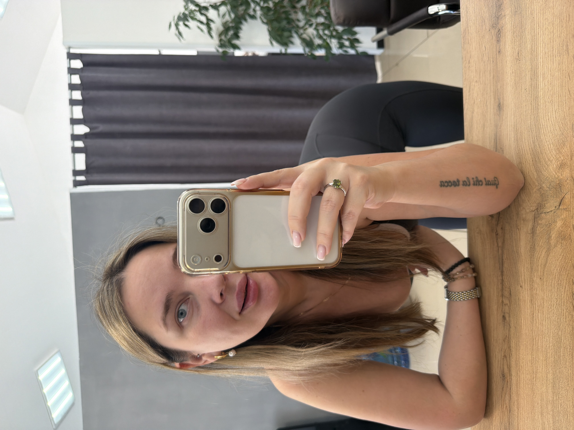 An indoor mirror selfie captures a woman holding a smartphone, reflecting her image. She has long, light brown hair, blue eyes, and fair skin, with a neutral expression as she looks into the phone's camera. Her nails feature a French manicure style. She wears a silver-colored ring with a green square-cut gemstone on her left ring finger. Her right arm rests on a light brown wooden surface, adorned with several bracelets, including beaded ones and a metallic mesh-style watch or bracelet. A small hoop earring with a pearl is visible on her left ear.
The smartphone, a light-colored model within a clear case, is held by her left hand. In the background reflection, dark horizontal window blinds are visible, along with parts of a green leafy plant and a dark piece of furniture, possibly a chair. The ceiling reflects bright fluorescent lighting panels. A tattoo in script is visible on her left forearm, which, when oriented correctly, reads "qual chi la tocca," an Italian phrase. The overall setting suggests a well-lit indoor environment during the day.