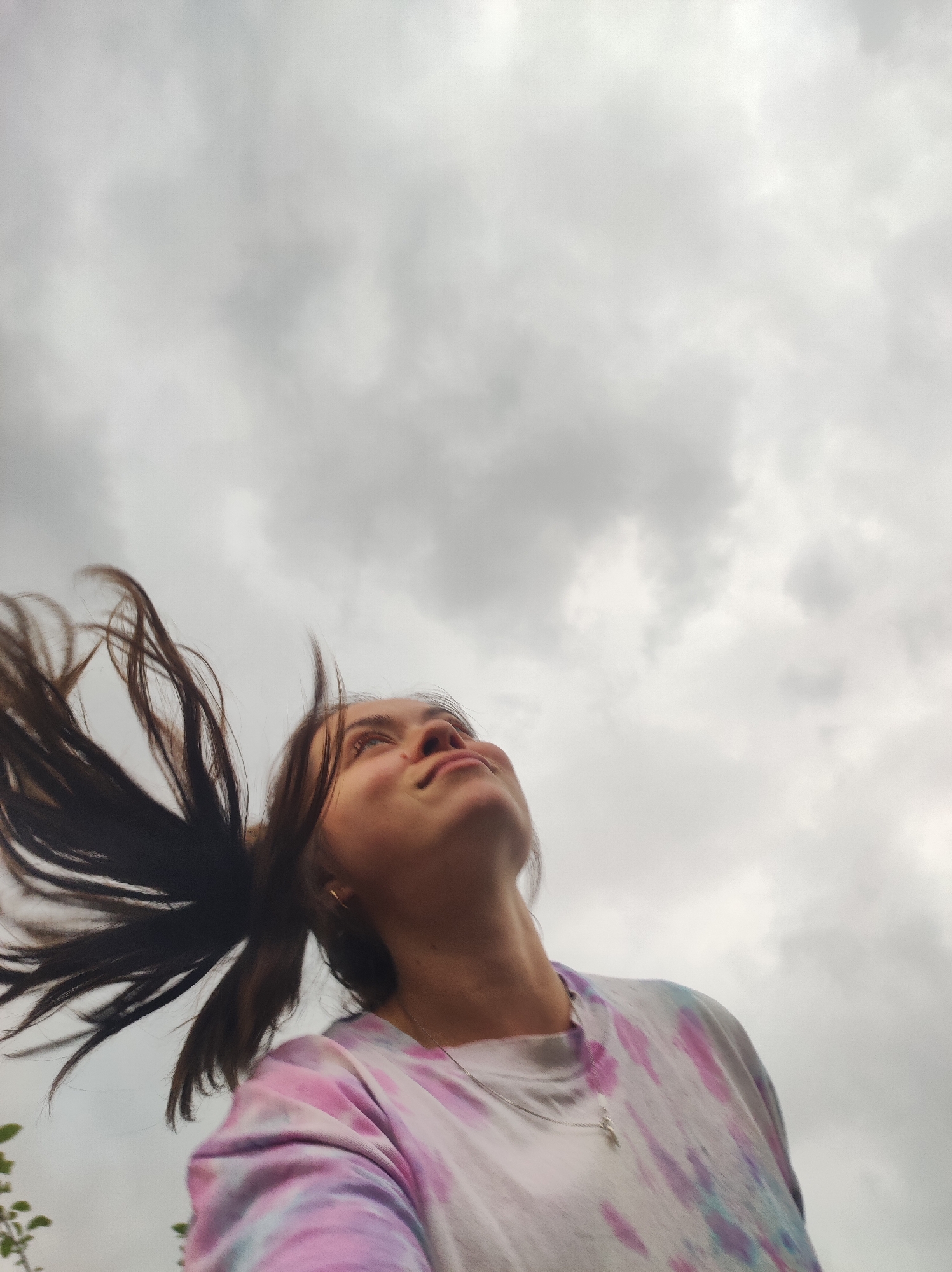 A young woman with dark hair, which is blowing in the wind, looks up at a cloudy sky. She is wearing a tie-dye t-shirt with pink, purple, and blue colors and a silver necklace with a cross pendant. The background consists of overcast, gray clouds, suggesting a potentially overcast or stormy day. The image appears to be taken from a low angle, looking upwards towards the woman and the sky. The location is not specified in the image.