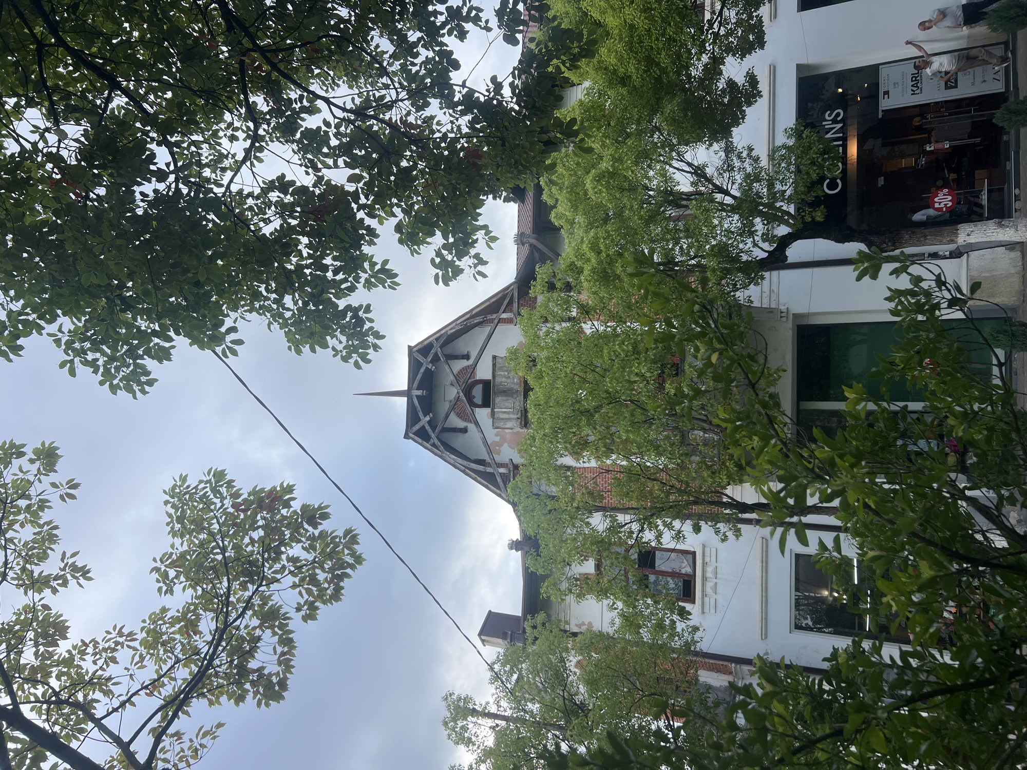 A building with a white facade and red brick accents is partially obscured by lush green trees. The building features a steep, gabled roof with exposed wooden beams and a spire at its peak. Several windows are visible, some appearing to be in disrepair. A storefront with the name "COLIN'S" is located on the ground floor, displaying clothing and a "-50%" sign. Two men are standing outside the storefront, with one gesturing upwards and the other looking towards the store. The sky above is overcast.