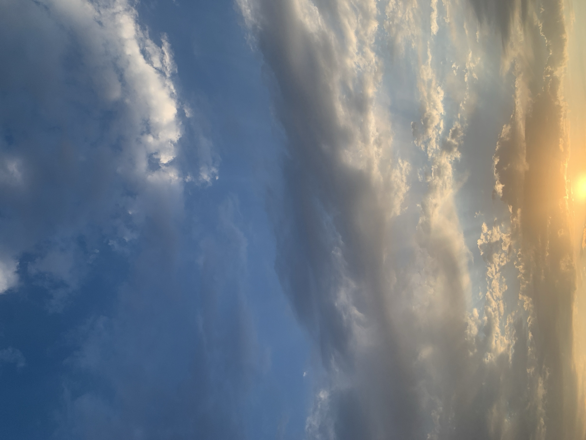 The image captures a sky view over Komsomolskoe, Kyrgyzstan, during what appears to be sunset. The sky is filled with a dynamic array of clouds, ranging from light and wispy to darker, more substantial formations. Sunlight is breaking through some of the clouds, creating visible rays of light that illuminate the scene. The lower portion of the image is dominated by the warm, golden hues of the setting sun, casting a soft glow. The overall impression is one of natural beauty and the dramatic transition of light as day turns into evening. No people, subjects, or specific objects are visible within the frame, only the sky and its cloud formations.