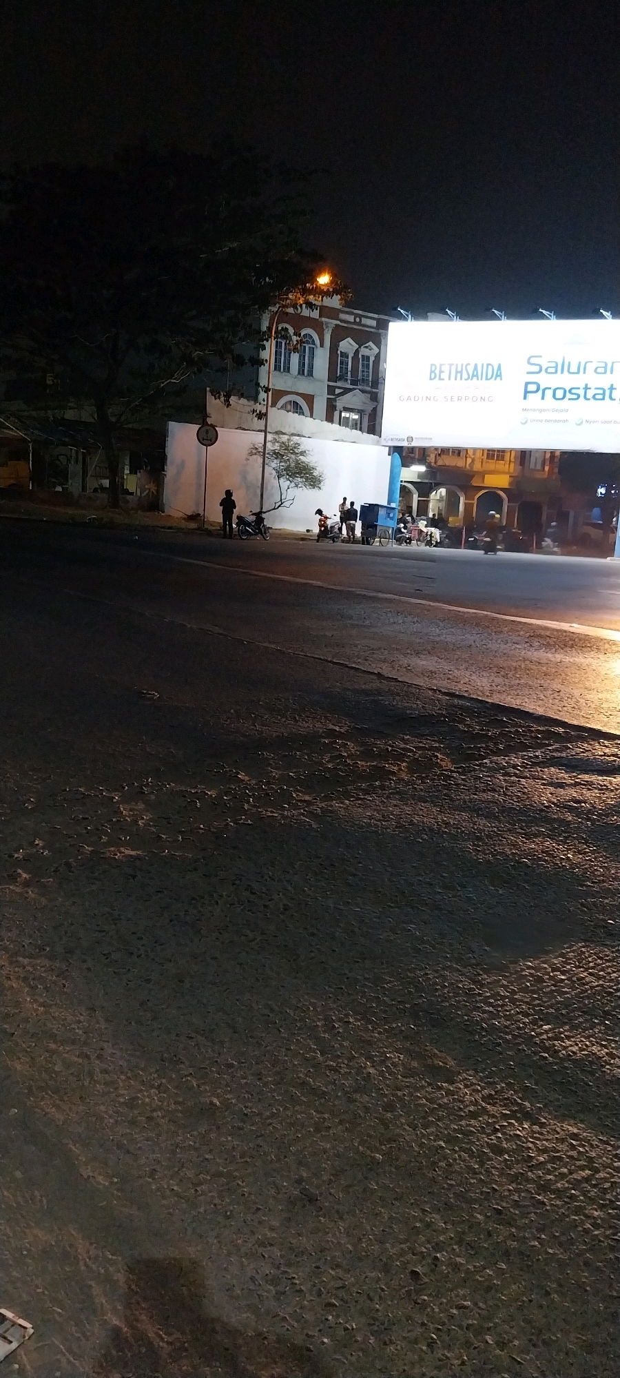 The image depicts a street scene at night in Kelapa Dua, Indonesia. A large billboard advertising "BETHSIDA" and "Saluran Prostate" is illuminated on the right side of the frame. In front of the billboard, a building with distinct arched windows is visible. On the street, several individuals are gathered near parked motorcycles and a blue cart. A lone figure stands beside a motorcycle on the left side of the street. The road surface is asphalt with visible patches and textures. The environment is dimly lit, with the primary light sources appearing to be the billboard and streetlights.