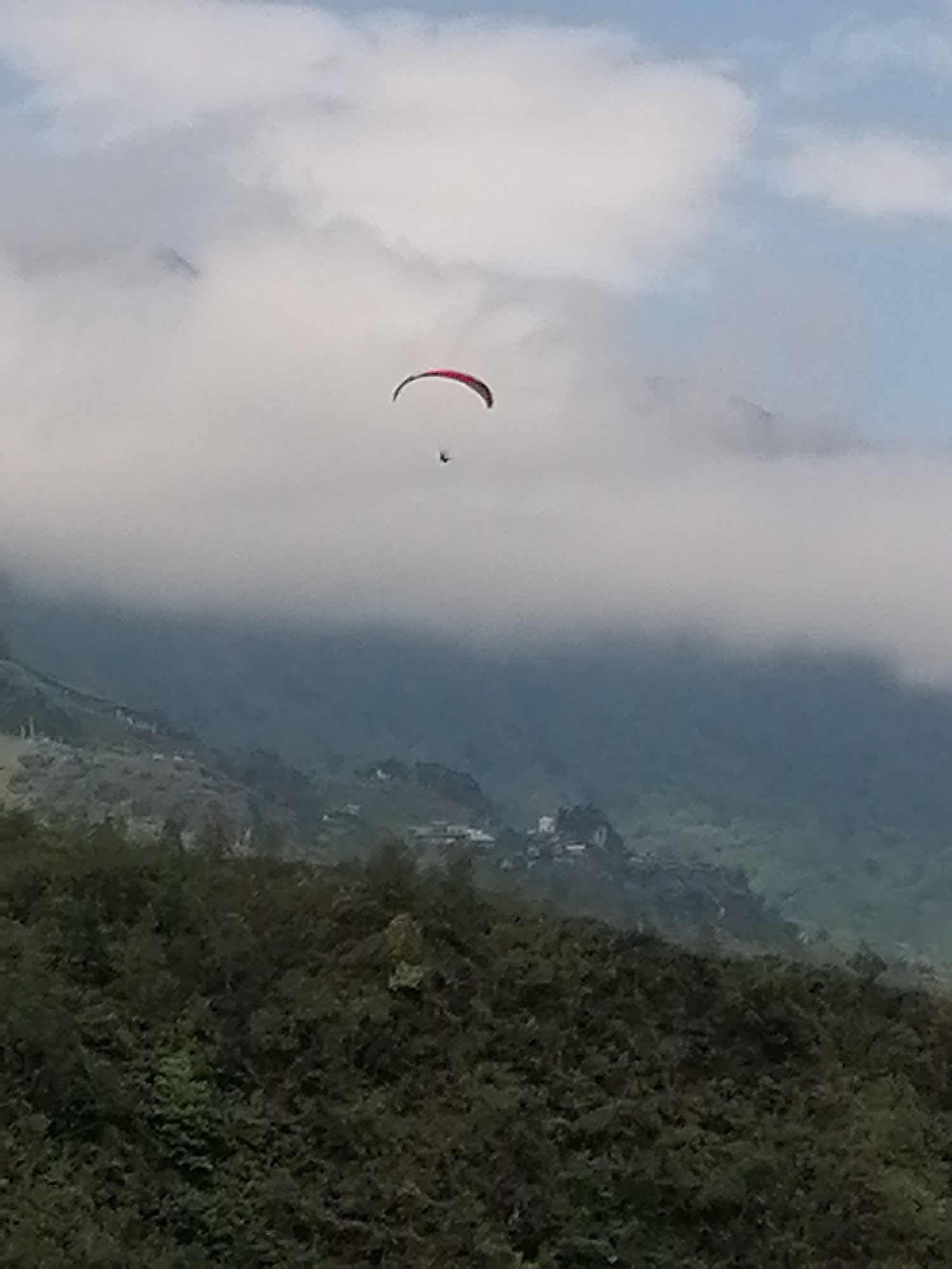 The image shows a paraglider flying in a hazy sky, likely in the mountainous region near Sa Pa Ward, Vietnam. The paraglider has a red and black canopy and a single person is suspended beneath it. The background consists of a lush, green mountain slope, partially obscured by low-lying clouds or fog. A small village or settlement is visible further up the mountain. The sky is a mix of pale blue and white, suggesting overcast conditions.