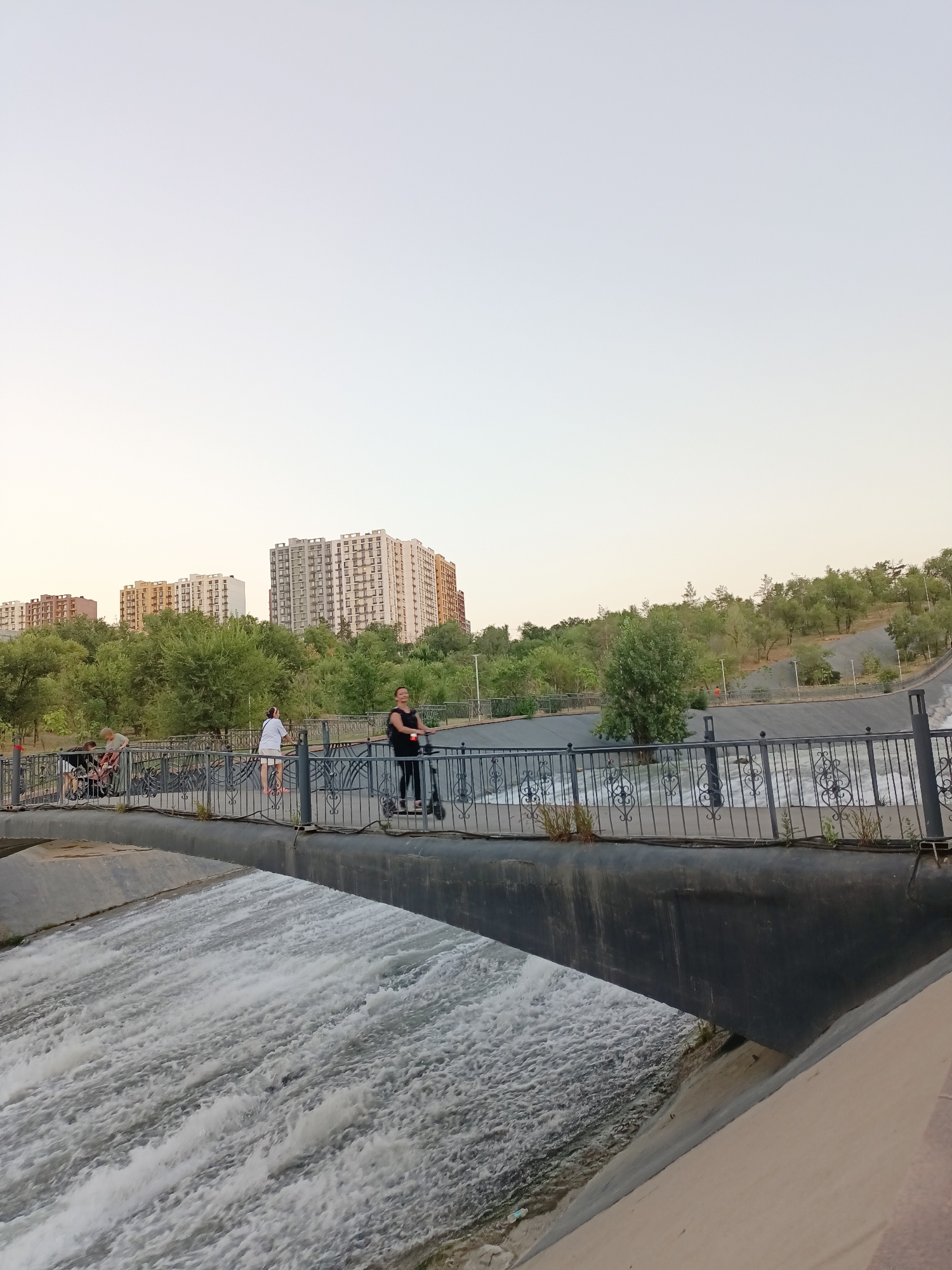 The image depicts a concrete canal with water flowing rapidly under a bridge. People are visible on the bridge, including a woman riding an electric scooter, two women walking, and a person pushing a stroller. Behind the bridge, there is lush greenery and several multi-story apartment buildings. The sky is clear and pale, suggesting it is either early morning or late evening. The overall scene is characteristic of a public park or recreational area within a city, likely Almaty, Kazakhstan, given the architecture and vegetation.