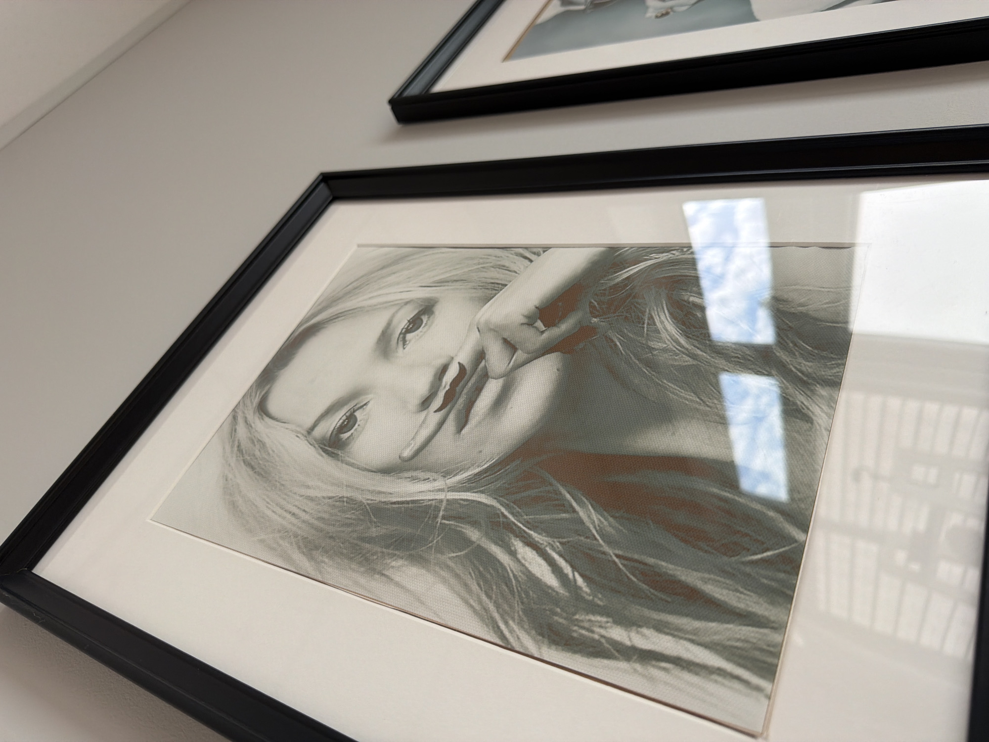 The image shows a close-up of two framed portraits hanging on a white wall. The portrait in the foreground is a black and white image of a woman with blonde hair, her finger raised to her mouth, creating a mustache effect. The print has a textured appearance, possibly a canvas or a fine mesh. Reflections of a window with blue sky and clouds are visible on the glass of the frame. The second portrait, partially visible to the right, is also framed in black and appears to be a portrait of a person. The scene is indoors and no specific location in Riga, Latvia, can be determined.