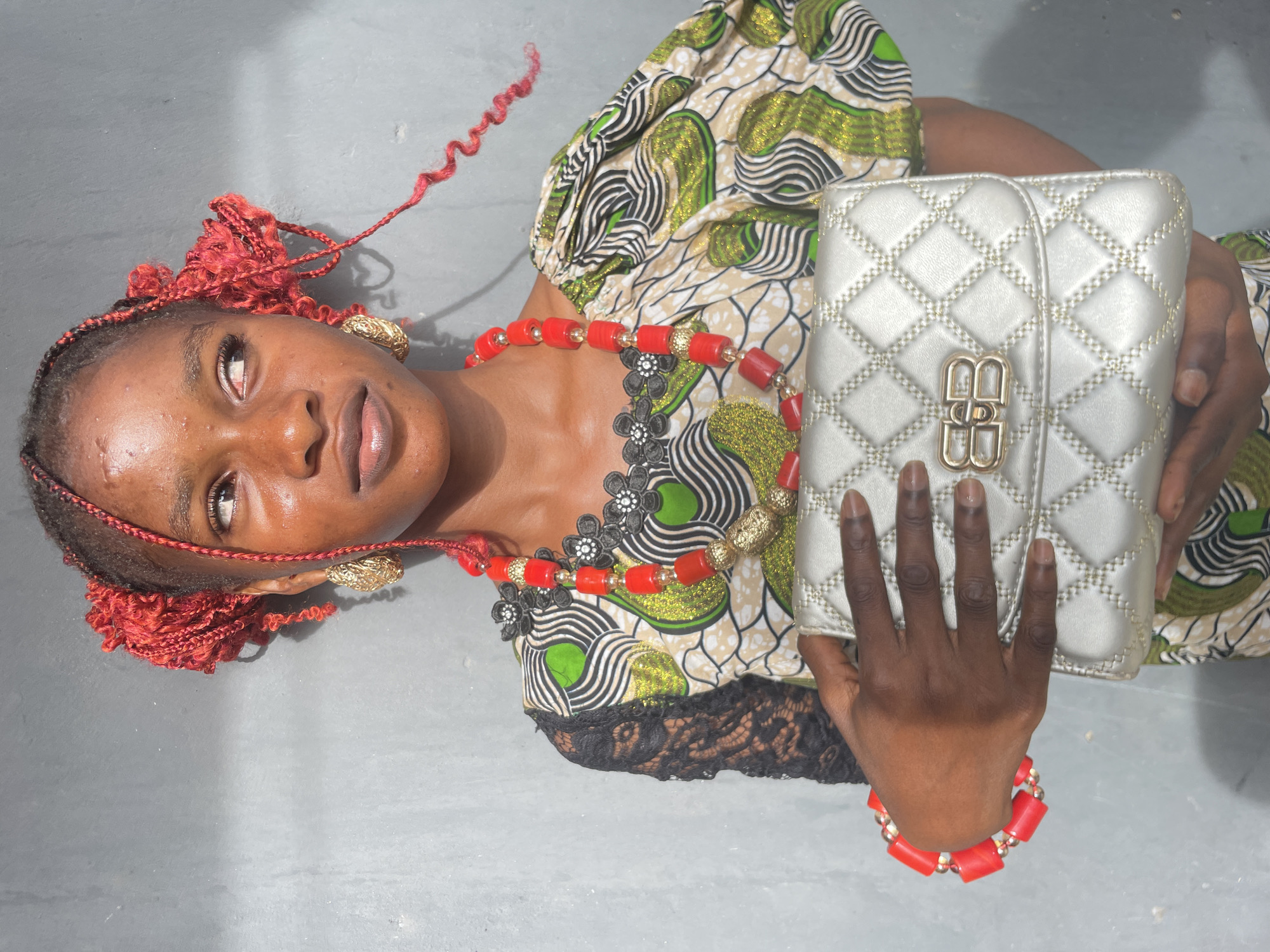 A person, likely a woman, is depicted lying on a light grey, possibly concrete, surface, gazing upwards and slightly to the left. Her dark skin has a noticeable sheen, reflecting bright natural light. Her hair is styled with vibrant red braids and curly extensions, some of which are loose and trail across the surface. She wears large, ornate gold earrings.
A chunky, multi-strand necklace made of bright red cylindrical beads and smaller gold spherical beads is prominent around her neck. The necklace also features black, flower-shaped appliqués with white centers. She is dressed in a short-sleeved top made of a patterned fabric, characterized by wavy lines and abstract shapes in shades of green, black, and white, with shimmering gold accents. A section of black lace trim is visible on the lower part of her torso.
Her left hand rests on a silver, quilted handbag, which has a distinct shiny gold clasp in the shape of a double "BB" logo. A matching red beaded bracelet is worn on her left wrist. The image captures her in a posed manner, holding the handbag. The lighting is bright, creating highlights and shadows, and the background is a plain, uniform grey. The setting is simple, potentially indoors or a shaded outdoor area in Badagry, Nigeria.