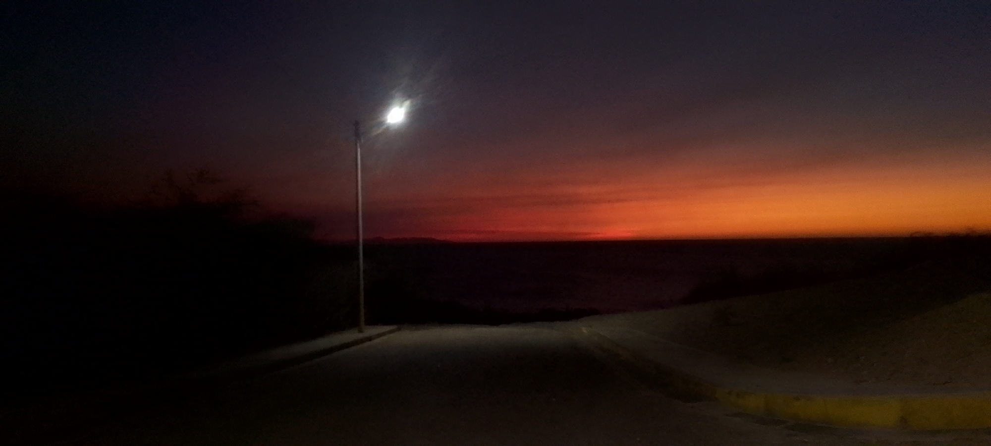 The image shows a street in Juan Griego, Venezuela at dusk. A streetlight illuminates the road, which slopes down towards a dark expanse of water. The sky is a gradient of deep blues and purples transitioning into shades of orange and red on the horizon. On the left, there is dark foliage, while the right side of the road is lined with a low curb. No people are visible.