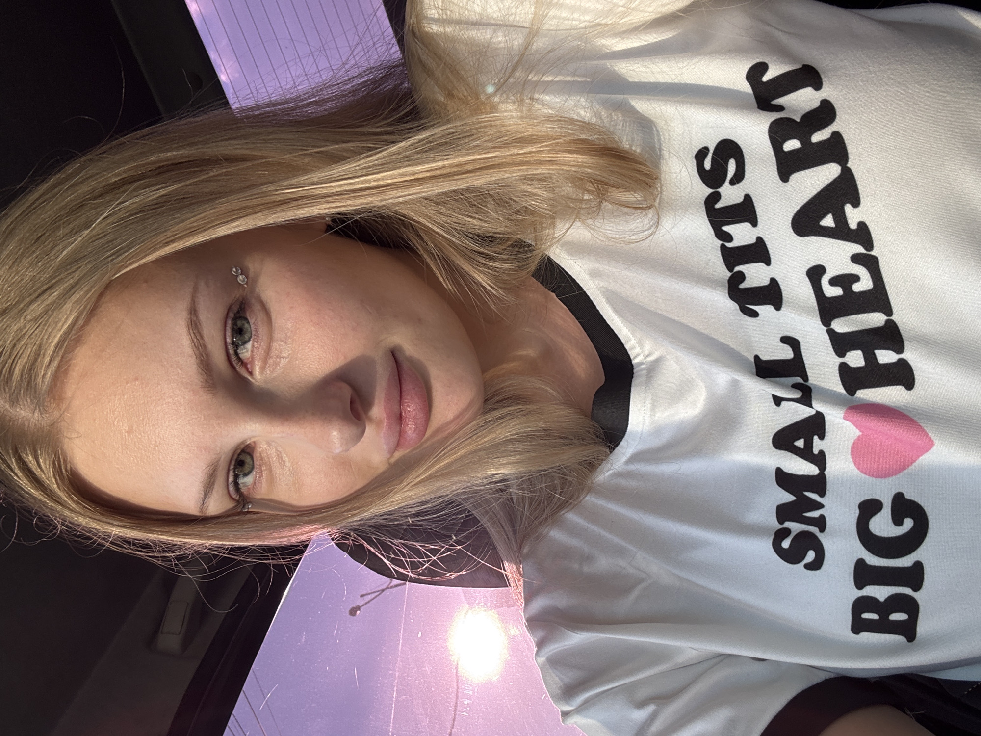 A young woman with blonde hair and light green eyes is taking a selfie in a car. She is wearing a white t-shirt with the words "SMALL TITS BIG HEART" and a pink heart printed on it. The sun is shining through the window behind her, creating a lens flare and a purple hue on the window. The image is taken from the chest up, and the background is blurred. The location is the city of Perm, Russia.