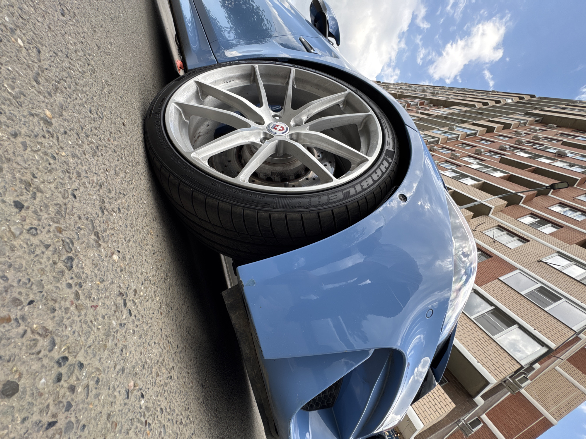 A low-angle, full shot shows the front quarter of a blue BMW M3 parked on asphalt. The car's front wheel, a silver multi-spoke alloy with a black tire, is prominently featured. The tire has white lettering that reads "HANKOOK VENTUS S1 EVO2". Behind the car, a multi-story brick apartment building dominates the background. The building has numerous windows and balconies, with several air conditioning units visible on the facade. The sky above is blue with scattered white clouds. A lamppost is attached to the building. The scene appears to be a typical urban street in Krasnodar, Russia, on a bright, possibly overcast day. There are no people or animals visible in the frame. The car's paint shows some minor scuffs on the lower front bumper area.