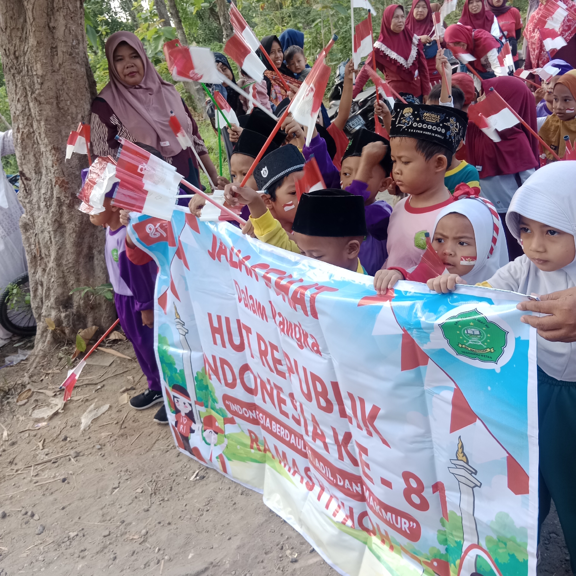 In Tegowanu Kulon, Indonesia, a group of children, some wearing traditional Islamic caps, are holding small Indonesian flags and a large white banner. The banner, which is partially unrolled, displays red text that reads "HUT REPUBLIK INDONESIA KE - 81" and "INDONESIA BERDAULAT, ADIL, DAN MAKMUR". A woman in a headscarf is visible in the background. The scene appears to be outdoors, with trees and dry ground contributing to the natural environment.