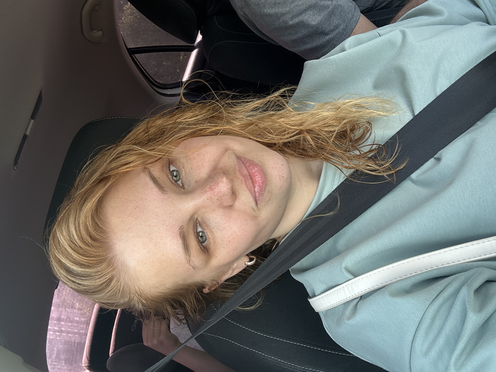 The image shows a young woman with blonde, wet hair and freckles, wearing a light blue t-shirt and a seatbelt. She is seated in the passenger seat of a car. The background of the image shows the interior of the car, including the headrest of the driver's seat and the window. The woman is looking at the camera with a slight smile. The location of Rusanovo, Russia, is not depicted in the image.