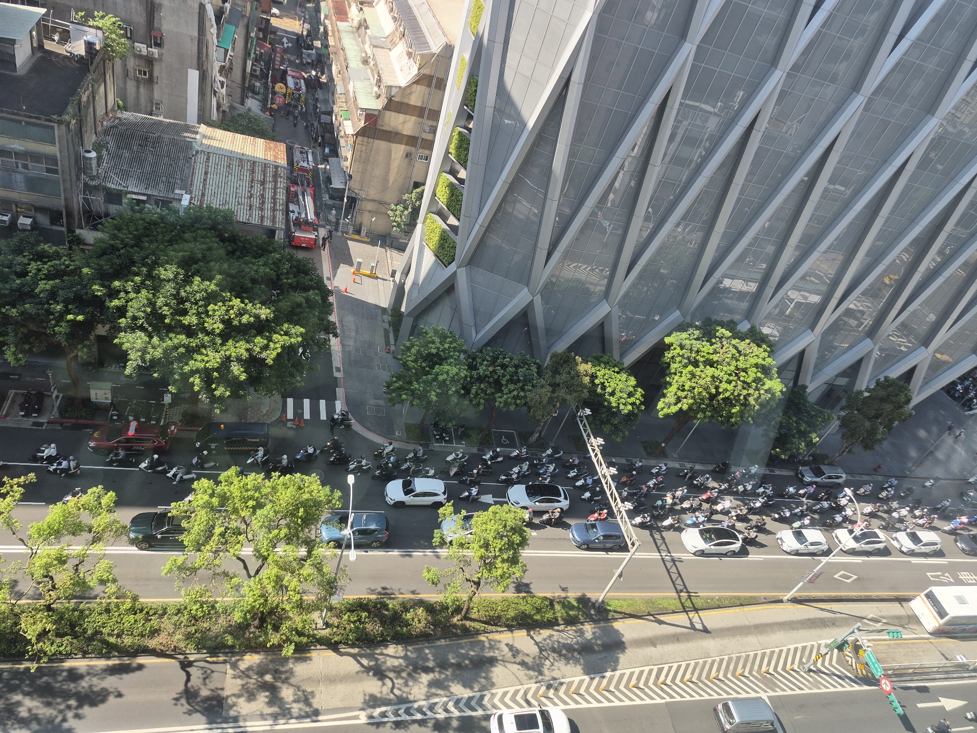 An aerial view of a street scene in Taipei, Taiwan, shows a busy intersection and a modern building. On the left, a narrow street is lined with older buildings and a few trees. In the center, several red fire trucks are parked, and people are gathered around them, possibly at an emergency scene. Further down the street, more vehicles and pedestrians are visible.
On the right, a large, contemporary building with a faceted, glass facade dominates the scene. Green planters are integrated into the building's design, adding a touch of nature. In front of the building, a wide street is filled with traffic, including numerous scooters and several cars. A traffic light and street signs are also present. The road is marked with white lines and dotted yellow lines, indicating lanes and traffic flow. Shadows from trees and buildings are cast across the road, suggesting it is a sunny day. The overall impression is a dynamic urban environment with a mix of old and new architecture, and constant movement of vehicles and people.