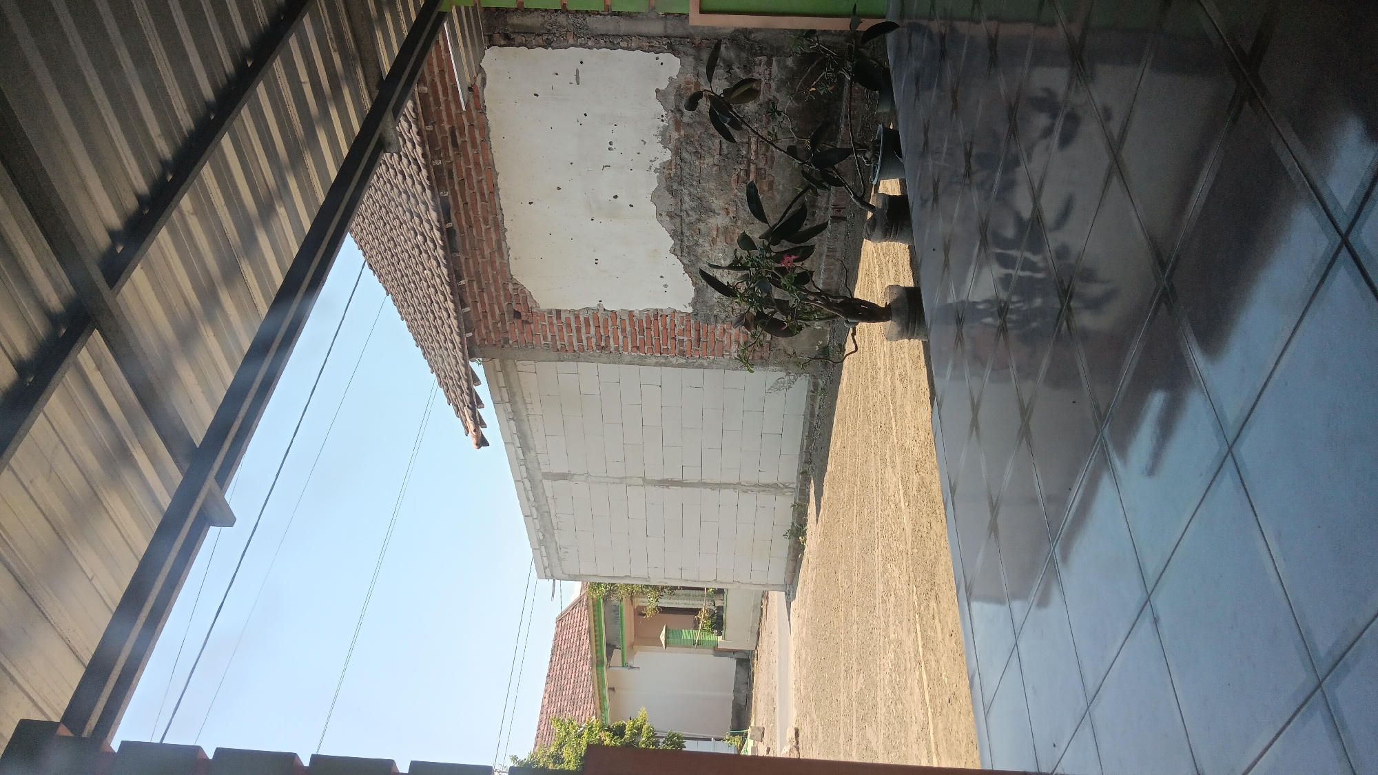 The image depicts a view from an elevated porch with a tiled floor, looking out onto a street scene in what is identified as the city of Sonobekel, Indonesia. The foreground is dominated by a reflective tiled floor. Beyond the porch, a dirt or gravel area is visible, leading to a street.
On the right side of the scene, a building under construction or in disrepair is present. It features a brick wall with areas of exposed brick and a large patch of peeling white plaster. Two potted plants with dark green leaves are situated in front of this wall. A dark, corrugated metal roof supported by a dark metal frame extends over the porch area, casting shadows.
To the left of the brick building, a new wall constructed of white concrete blocks is visible. Further in the background, a house with a red tiled roof and white walls is seen. A green structure, possibly a fence or decorative element, is also present in front of this house. Power lines are strung across the clear blue sky. No people are visible in the image. The overall impression is one of an outdoor, possibly residential or construction, area during daylight.
