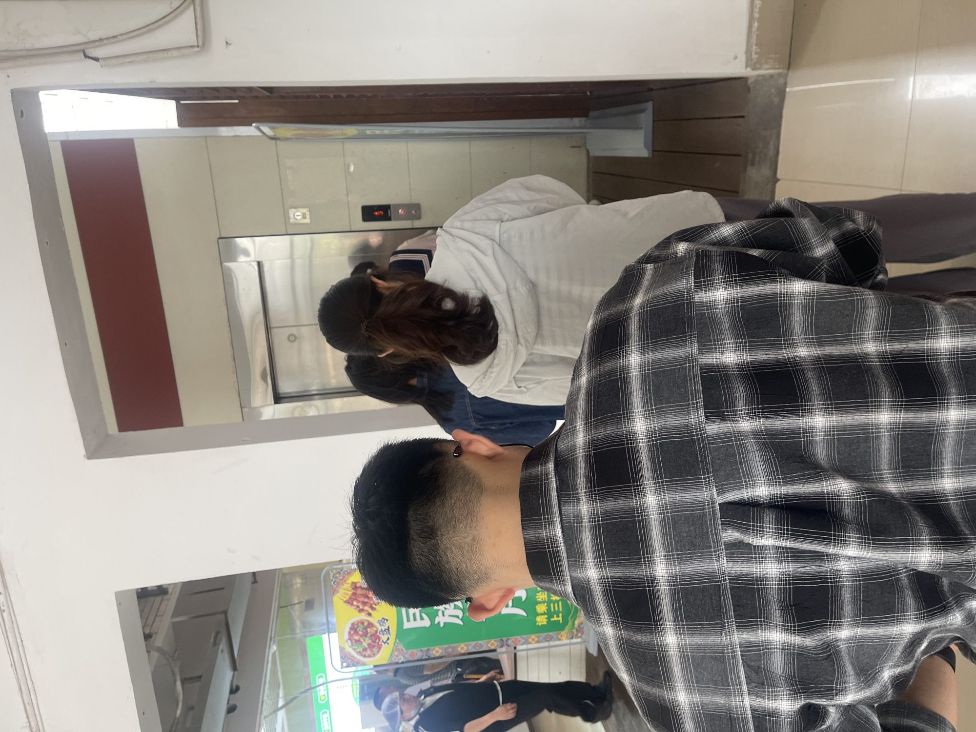 People are waiting near an elevator in a public space. The location is identified as Chongqing High-tech Zone, China. The foreground features a person in a plaid shirt, with other people in the background near the elevator. A menu sign is visible in the lower-left.
